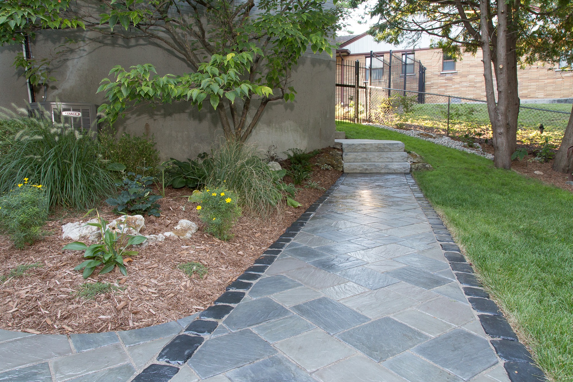 A neatly paved pathway winds through landscaped greenery, alongside a building, leading to a gate. Natural elements include trees and flowering plants.