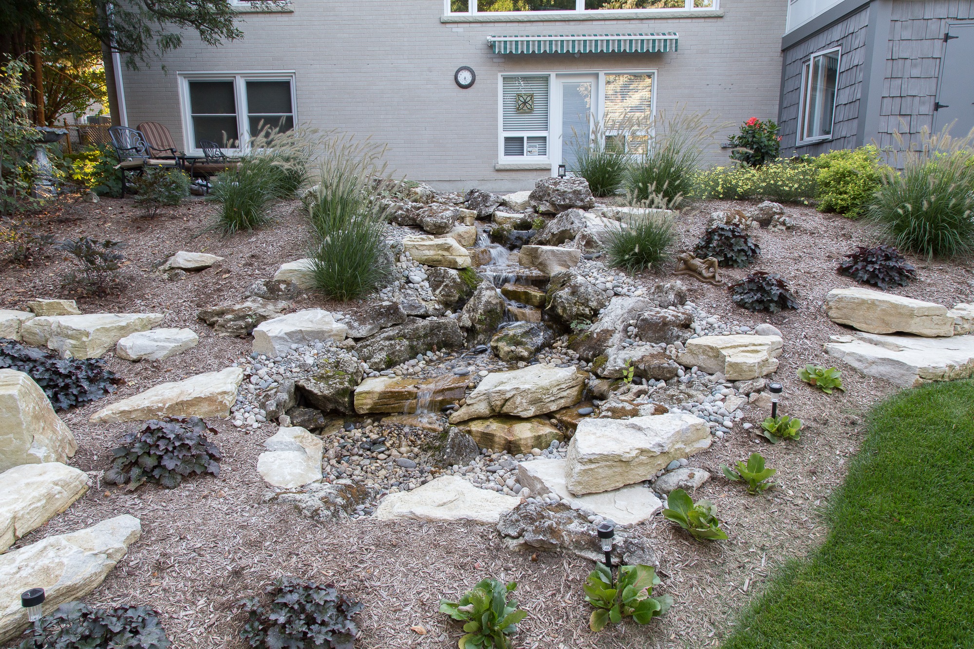 A landscaped backyard features a rocky stream, surrounded by plants and outdoor seating, adjacent to a light brick house with an awning.