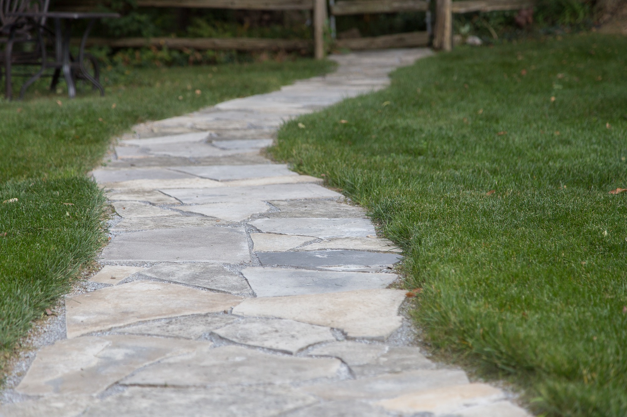 A stone pathway winds through a grassy garden area, bordered by a wooden fence with an empty metal chair set nearby.