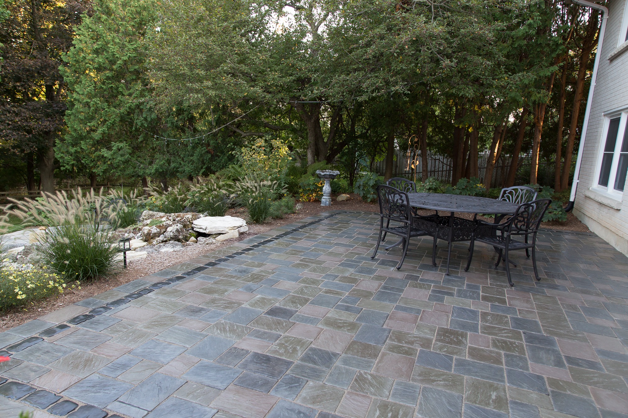 Spacious patio with multicolored stone tiles, metal table and chairs, surrounded by lush greenery and trees, creating a serene outdoor atmosphere.