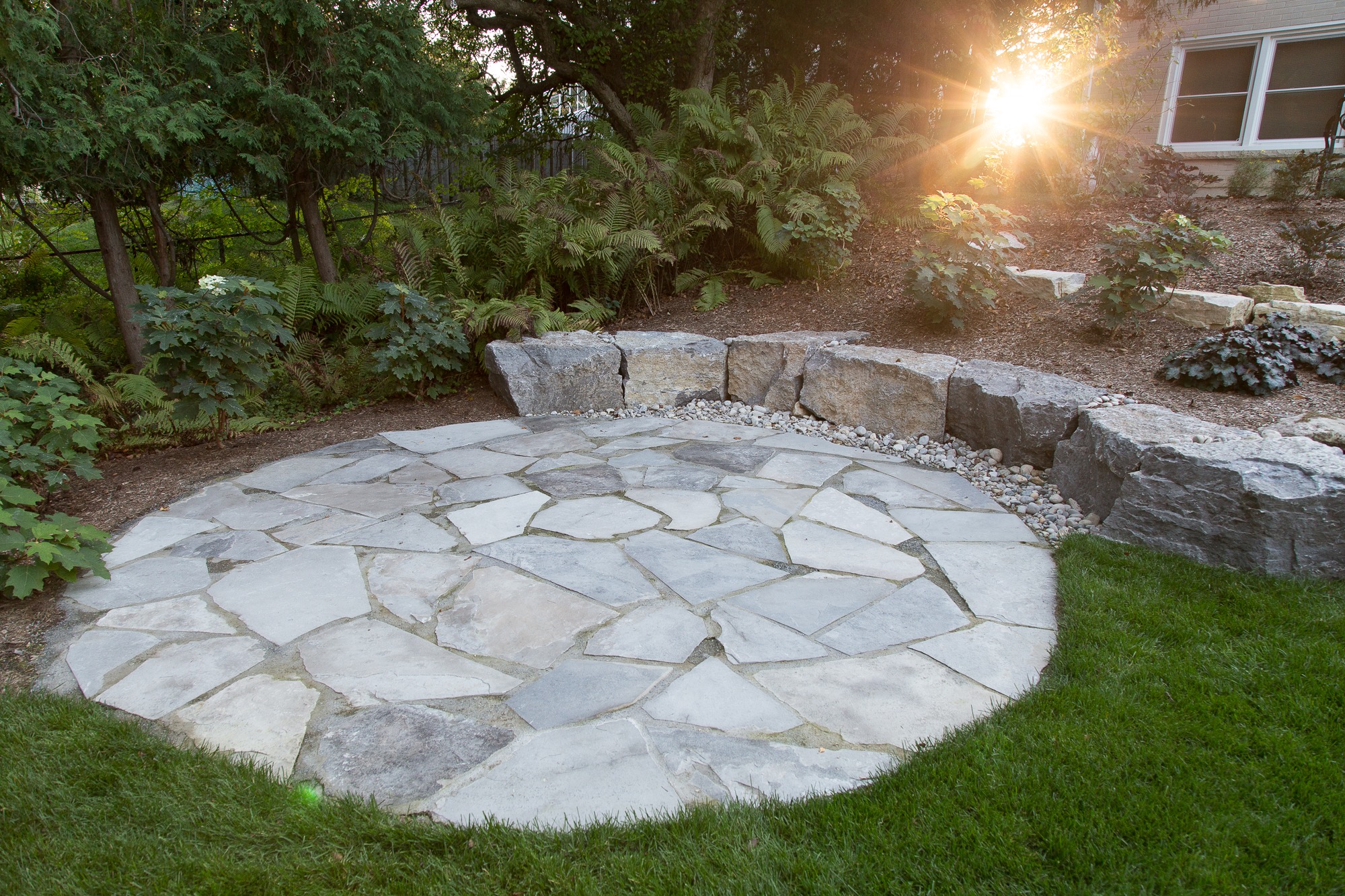 A circular stone patio surrounded by greenery, shrubs, and rocks, with sunlight peeking through trees, creating a serene garden atmosphere.