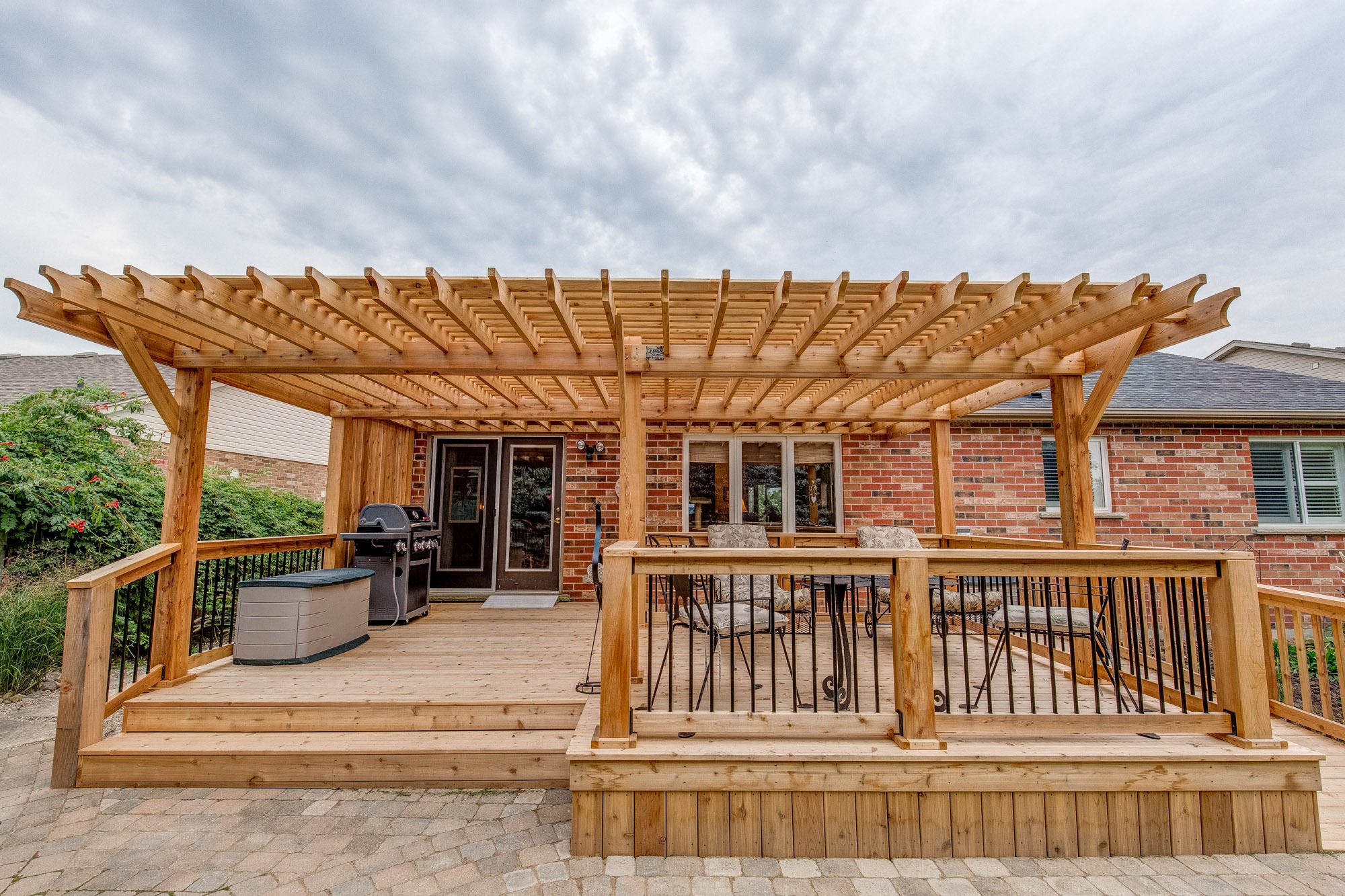 Wooden pergola over backyard deck with grill, patio furniture, and brick house. Overcast sky in the background adds a cozy ambiance.