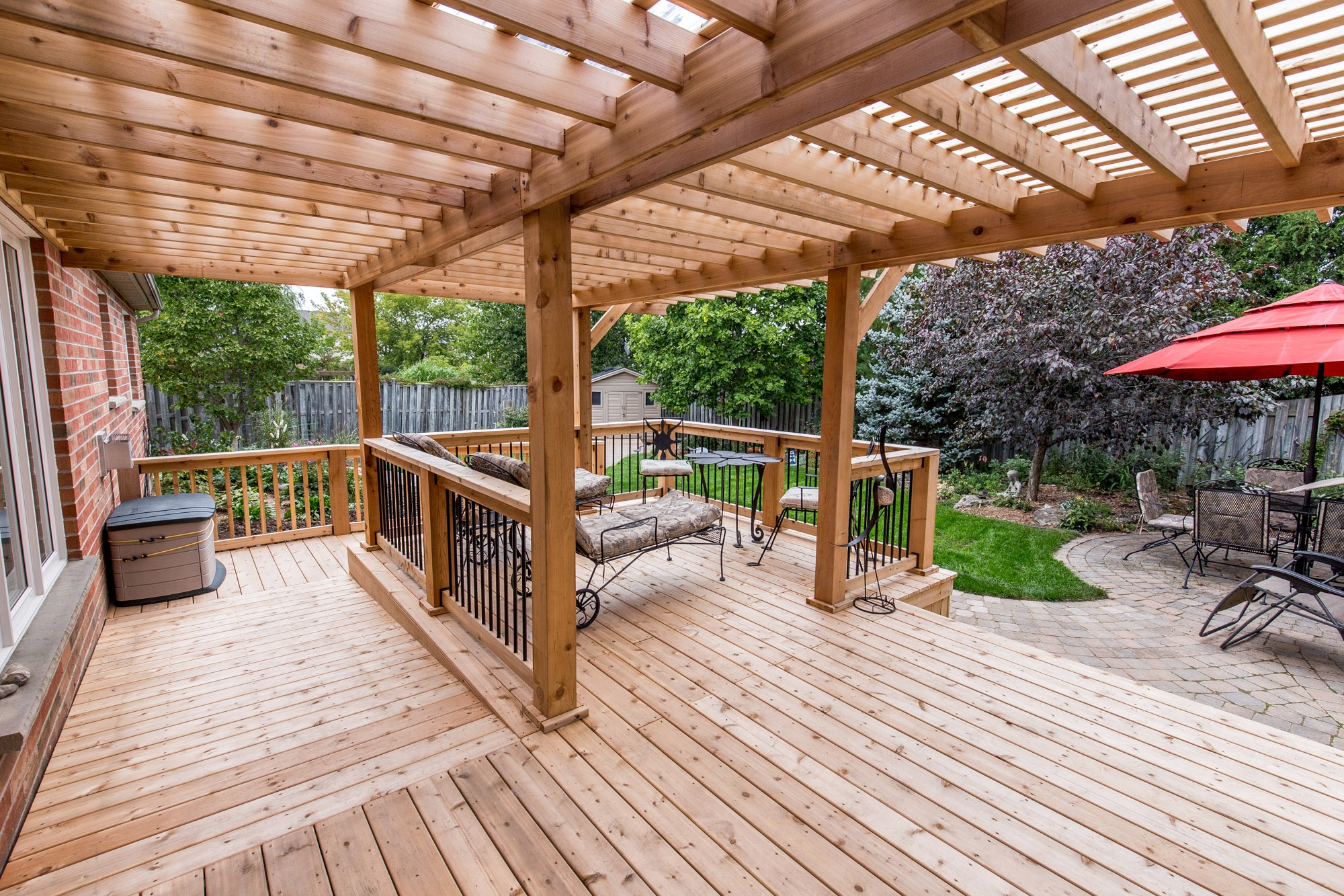 A wooden deck with pergola features patio furniture, red umbrella, and view of garden with trees and paved seating area.