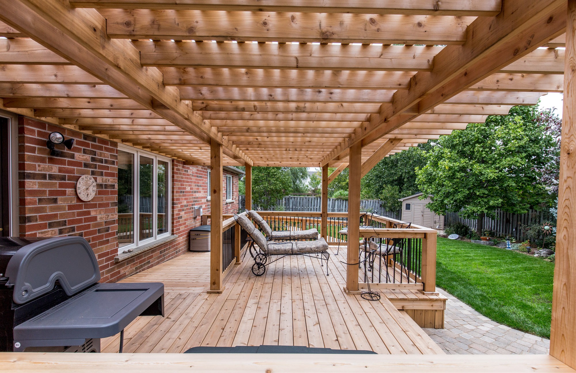 A wooden backyard deck with pergola, chaise lounges, grill, and lush green lawn. Surrounding fence and shed visible in the background.