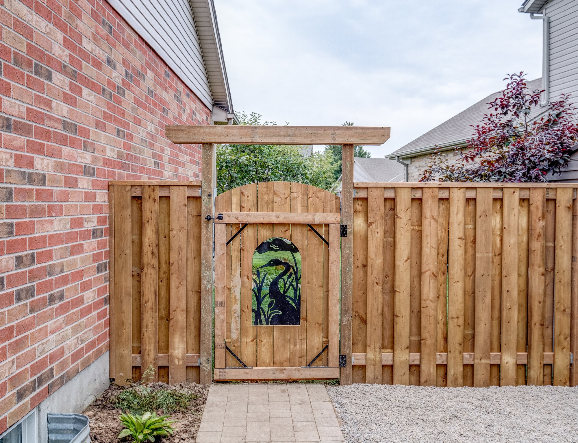 Wooden gate with painted design, nestled between houses. Brick and wooden fence create cozy ambiance, with small plants along the path. Overcast sky above.