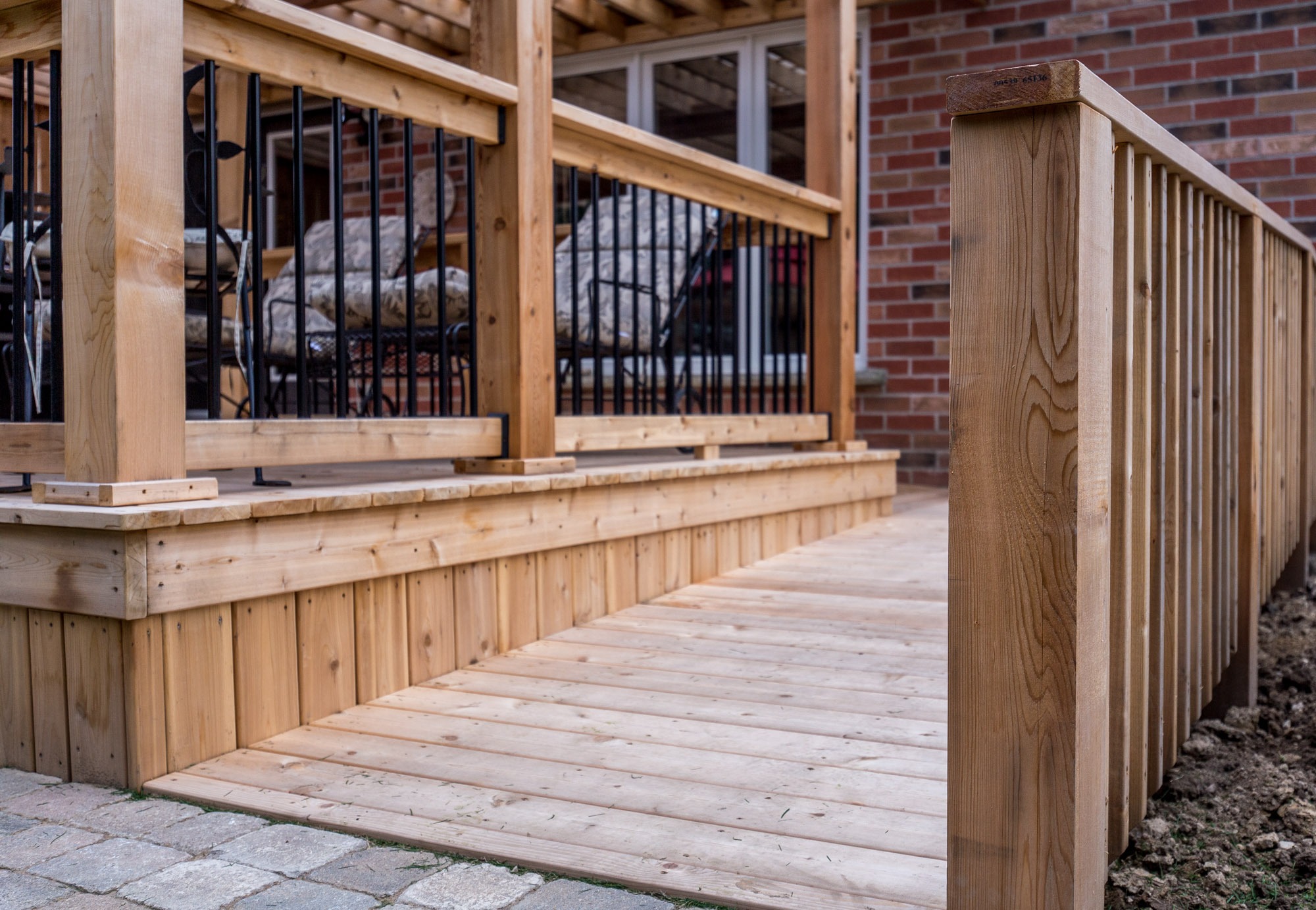 Wooden porch features railings, patio chairs, and a brick exterior. The pathway is made of light wood planks and leads to the entrance.