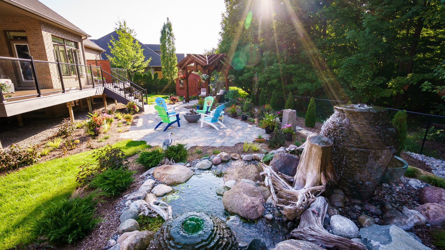 A tranquil backyard patio features colorful chairs, a fire pit, water feature, lush landscaping, and pergola, with sunlight streaming through trees.