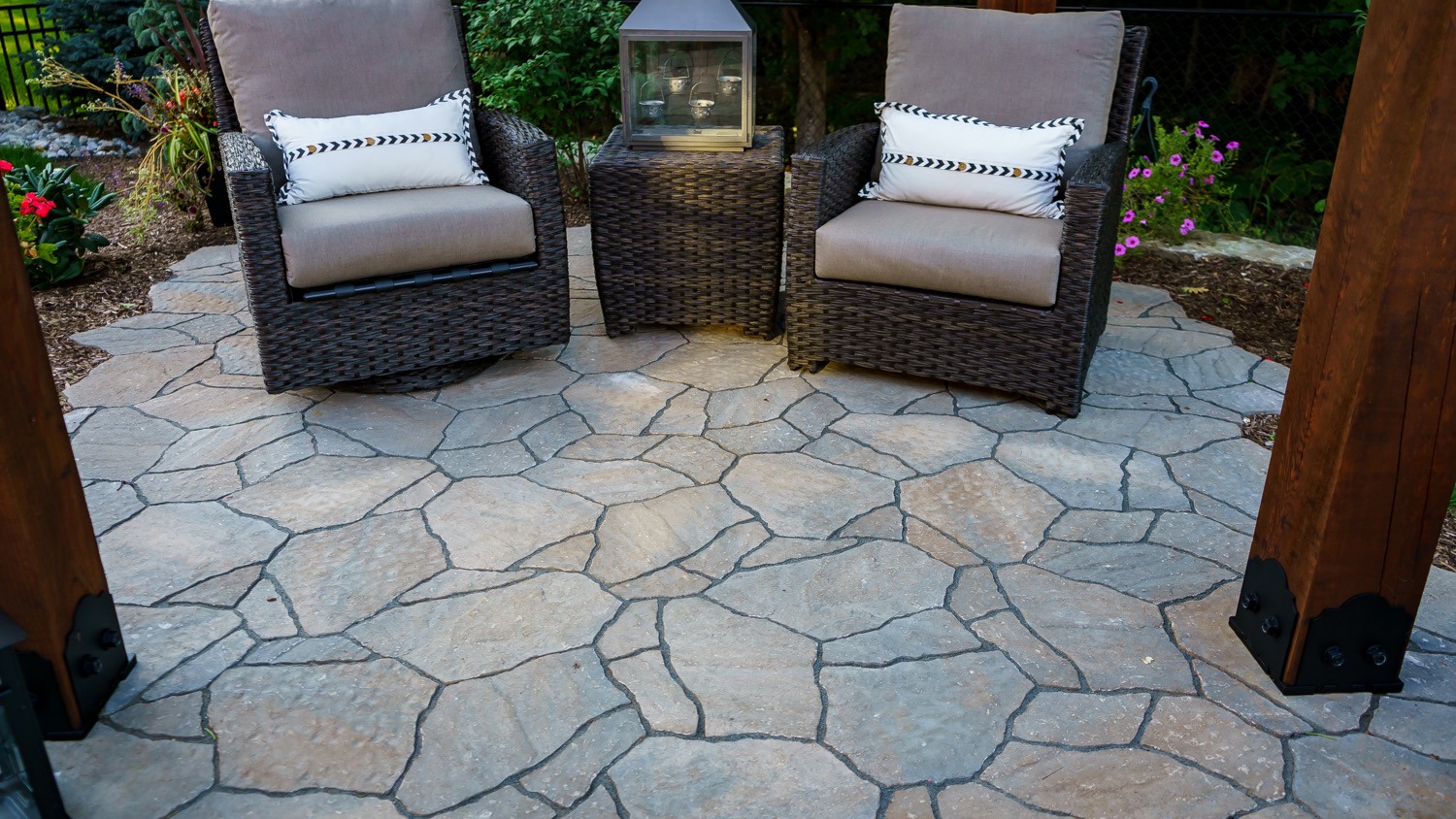 Outdoor patio with two cushioned chairs, small table, lantern, and surrounding greenery. Stone flooring adds an elegant touch to the cozy setting.