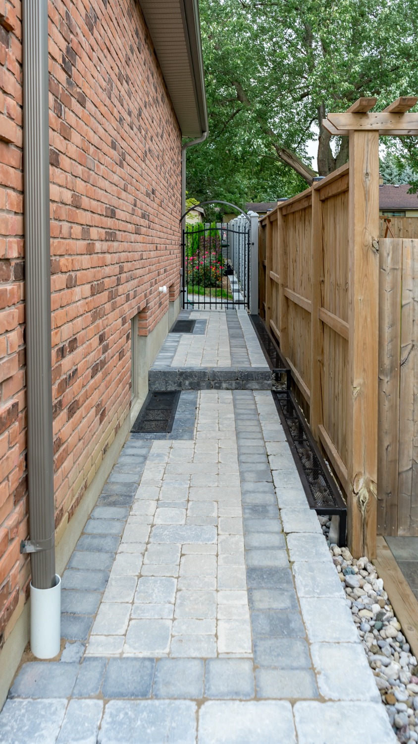 A narrow stone pathway runs between a brick wall and wooden fence, leading to a metal gate with plants and trees visible beyond.