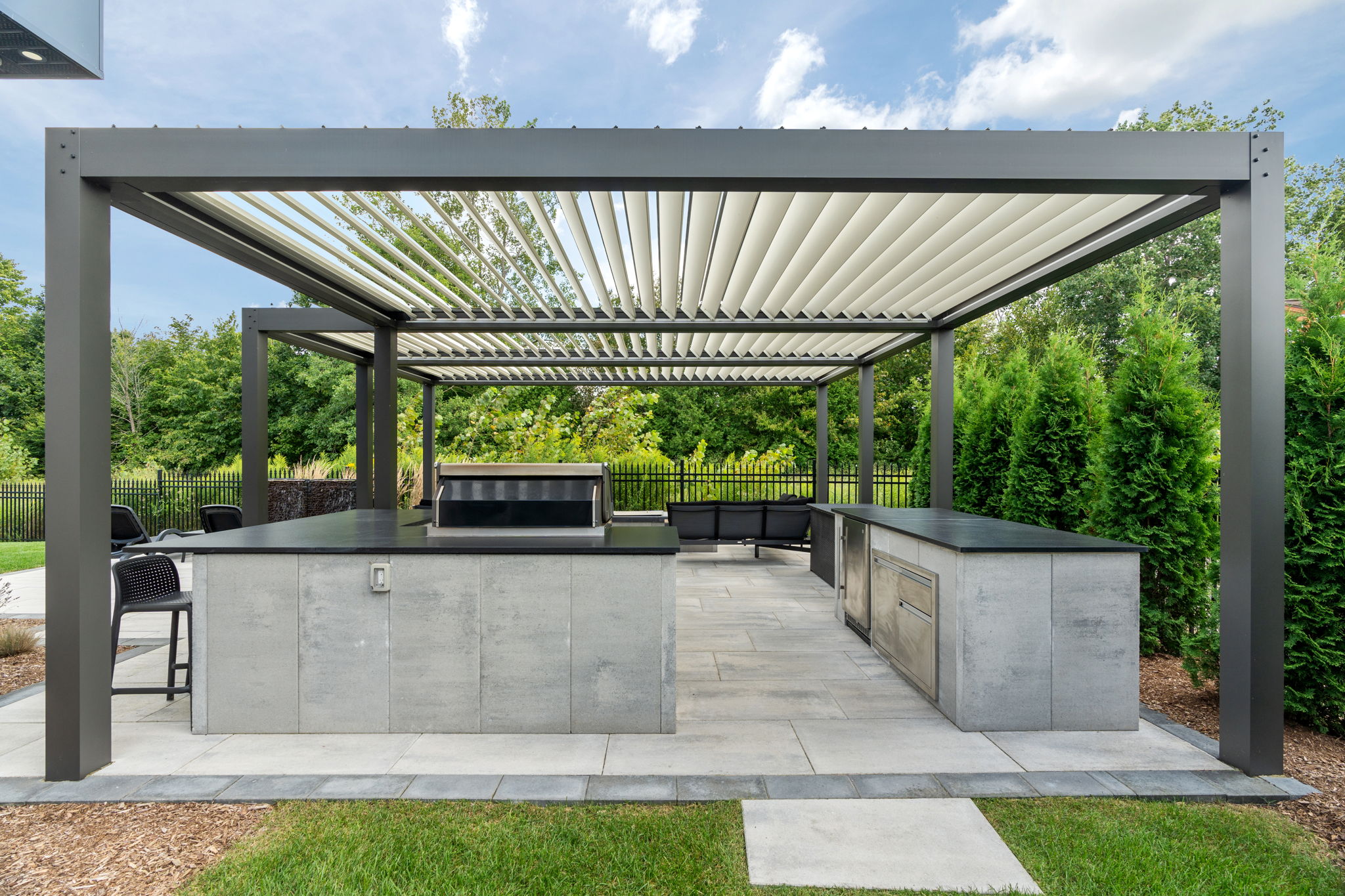 Modern outdoor kitchen with a sleek pergola, grill, and seating area. Surrounded by lush greenery and a neatly manicured lawn.