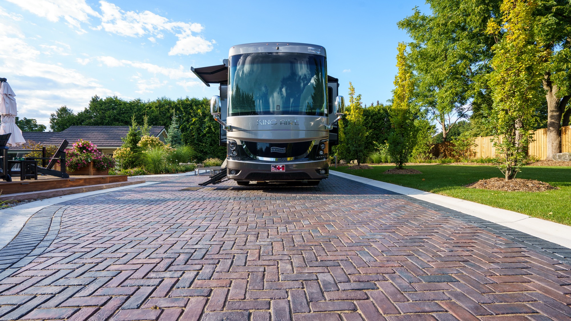 A luxurious motorhome parked on a brick driveway, surrounded by lush green trees and a neatly landscaped garden under a clear blue sky.