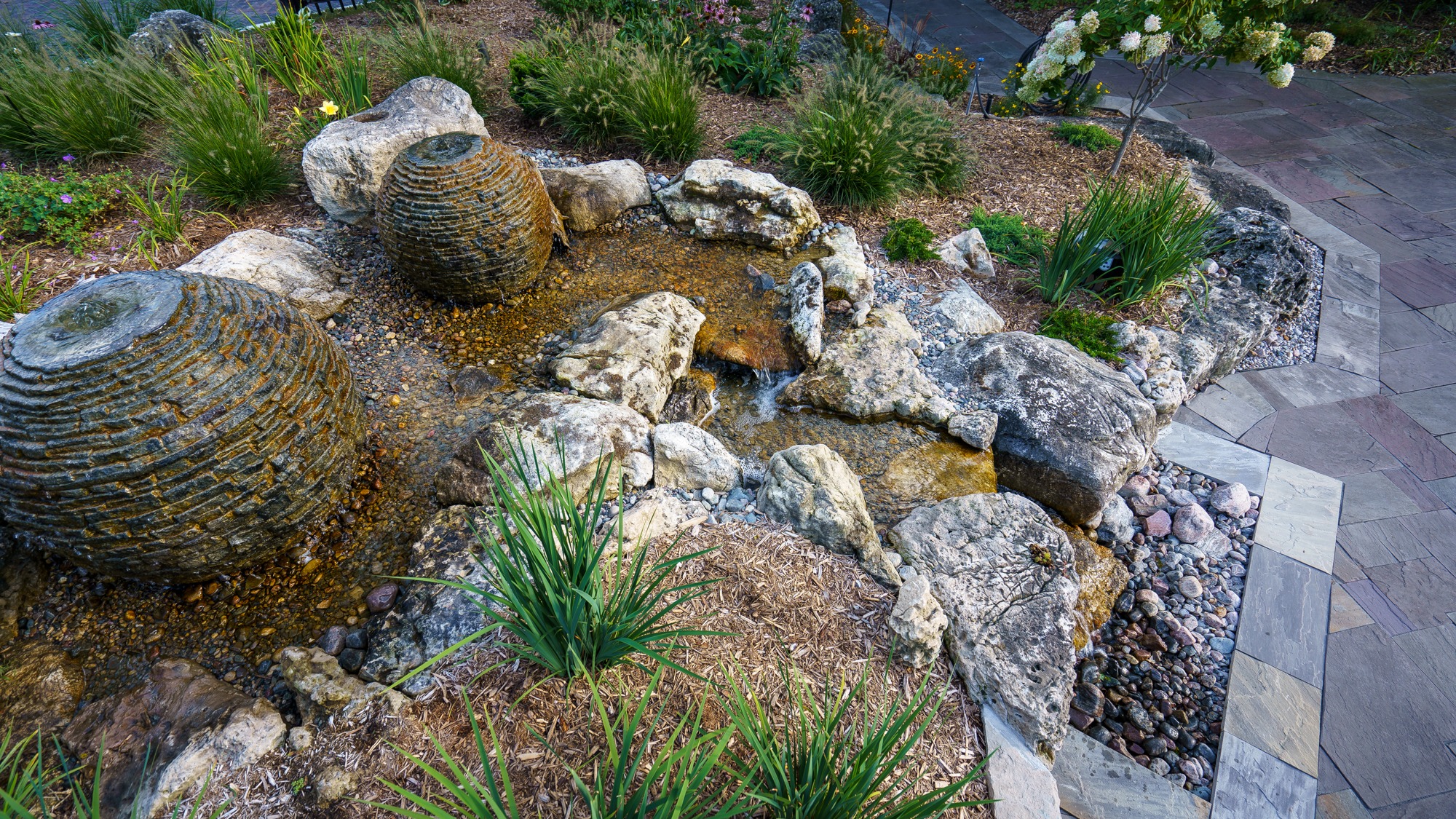 A serene garden features a small stone water feature with smooth stones, surrounded by lush greenery and a tiled walkway.
