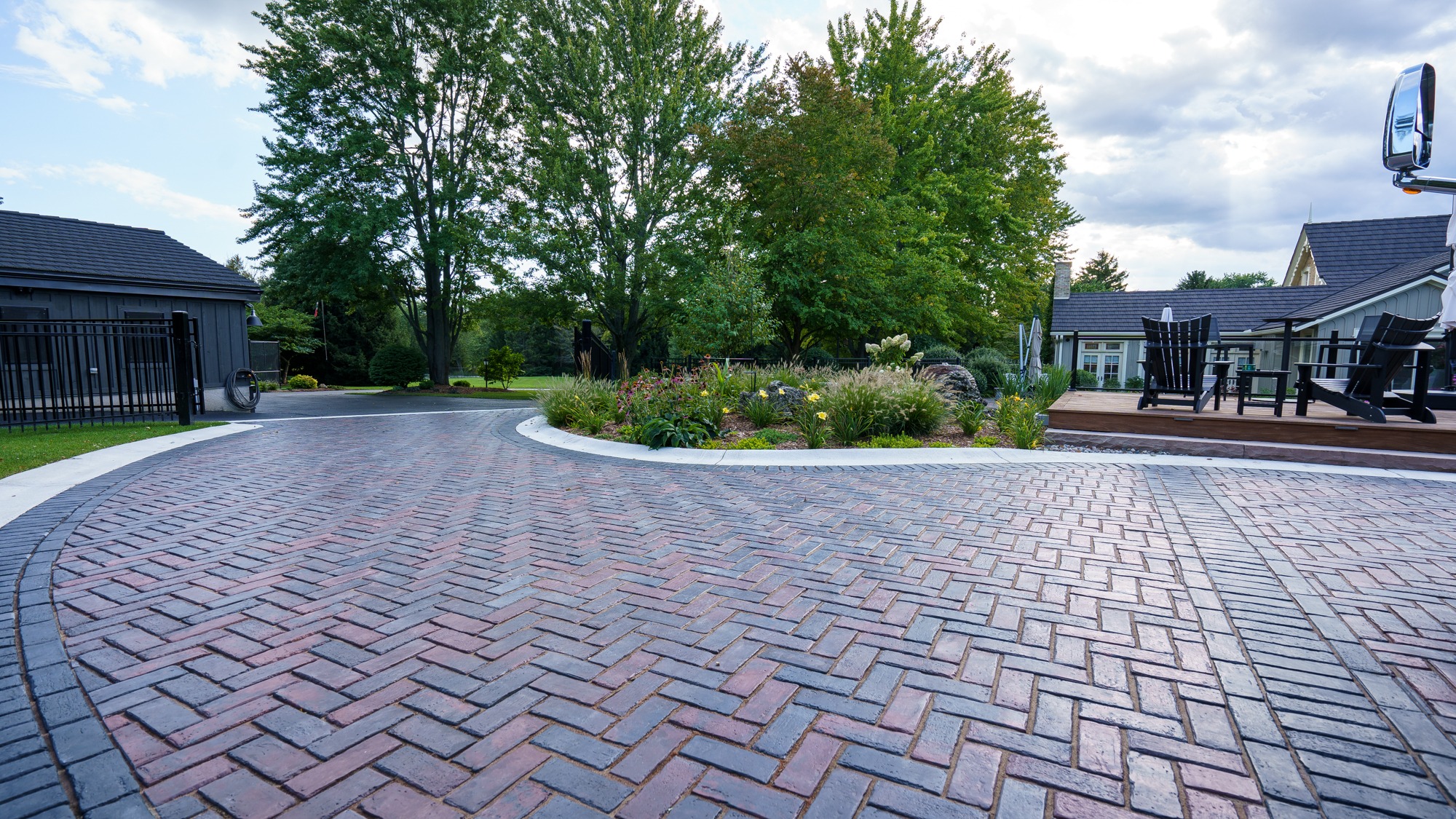 A brick-paved driveway with surrounding garden beds and outdoor seating area. Trees and modern buildings are visible in a serene setting.
