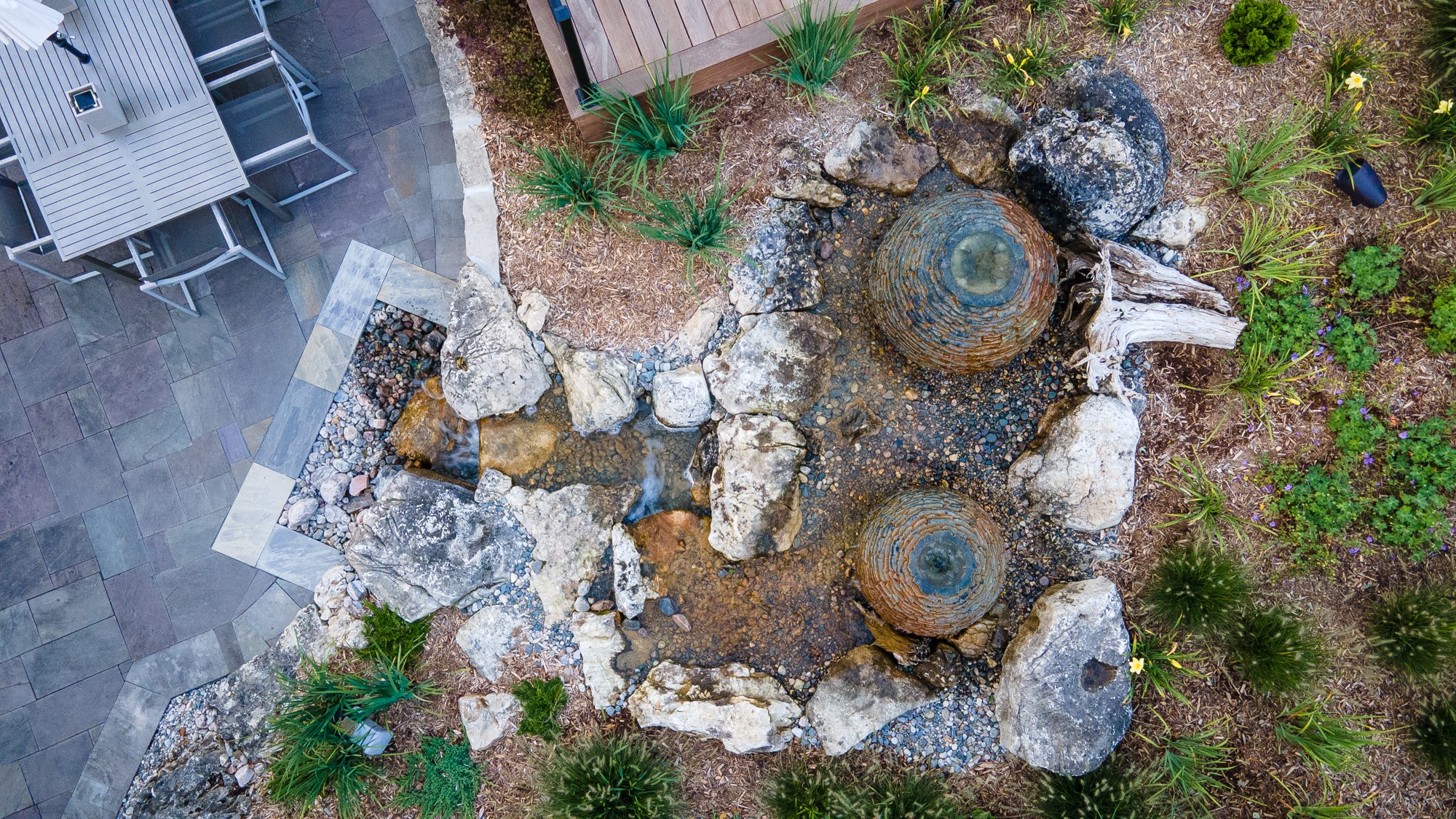 Aerial view of a landscaped rock garden with a small waterfall, adjacent to a tiled patio and table with chairs.