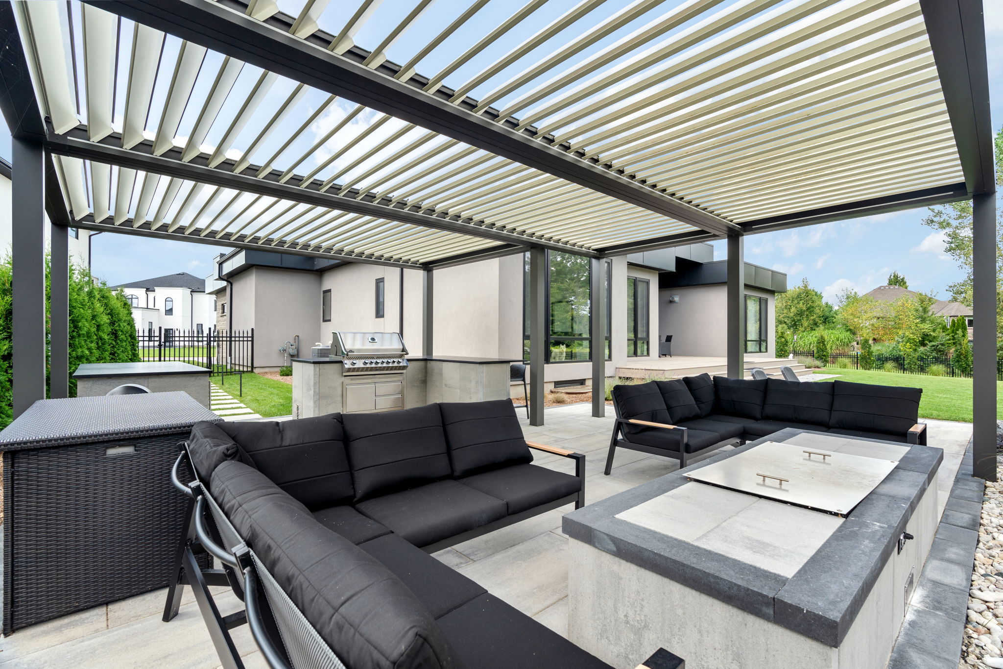 Modern outdoor patio with black furniture, pergola, grill, and lush greenery. Contemporary architectural design with sleek lines and minimalistic aesthetics. Peaceful setting.