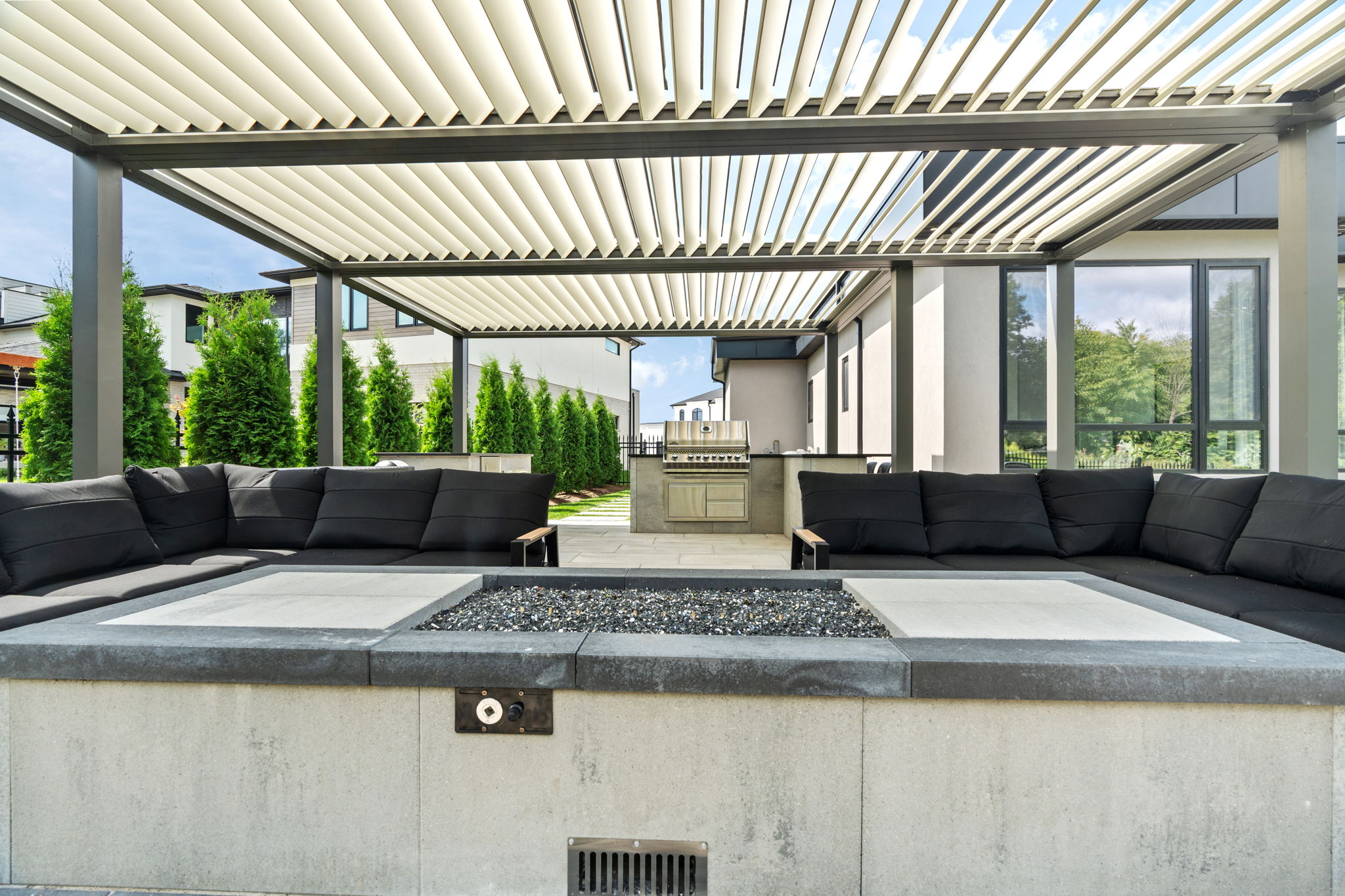 Modern patio with black sofas under a pergola, featuring a central fire pit and outdoor grill, surrounded by greenery and contemporary architecture.