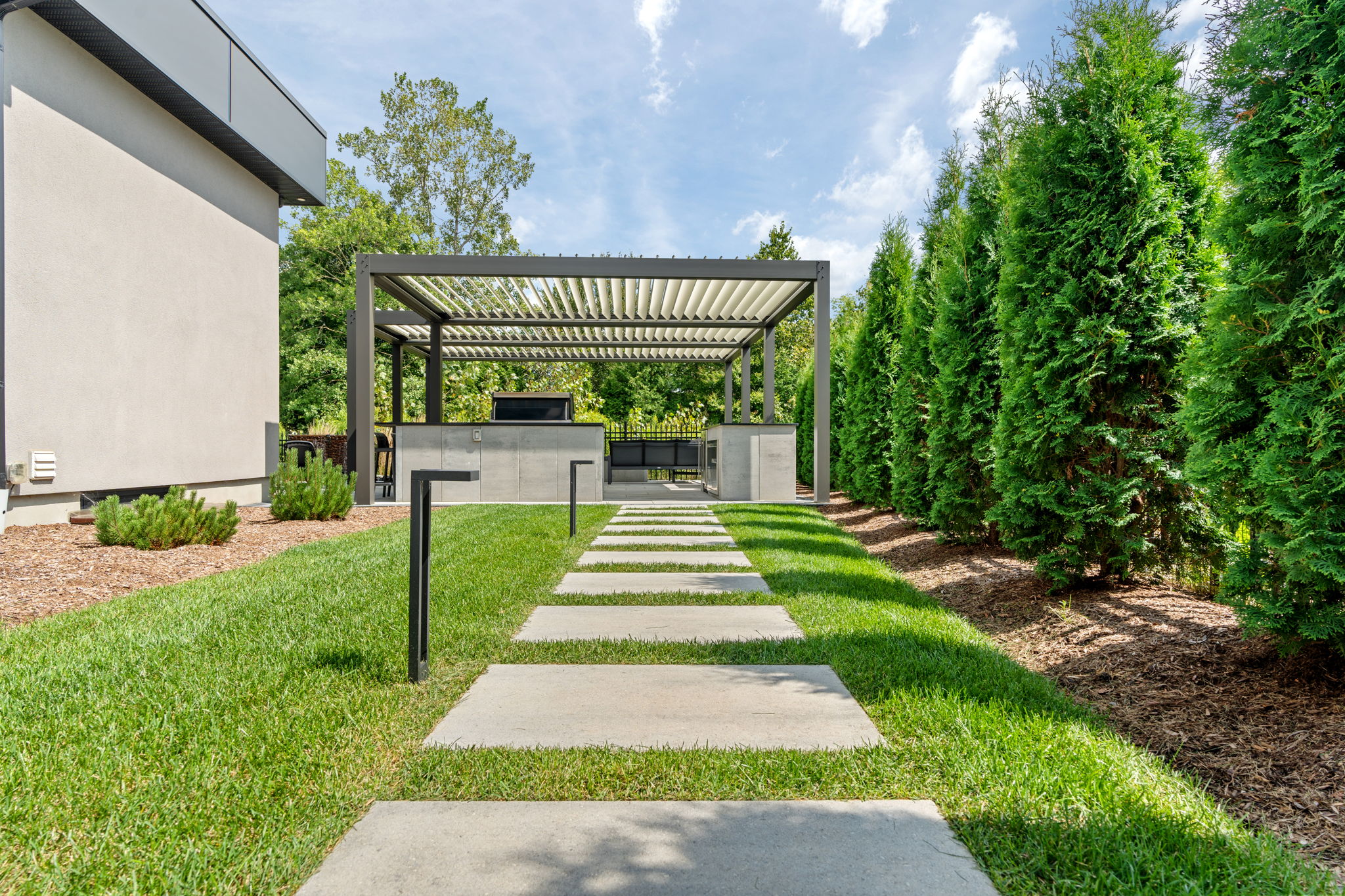 Modern backyard with a pergola, concrete path, neat lawn, and tall green hedges, set against a clear blue sky.