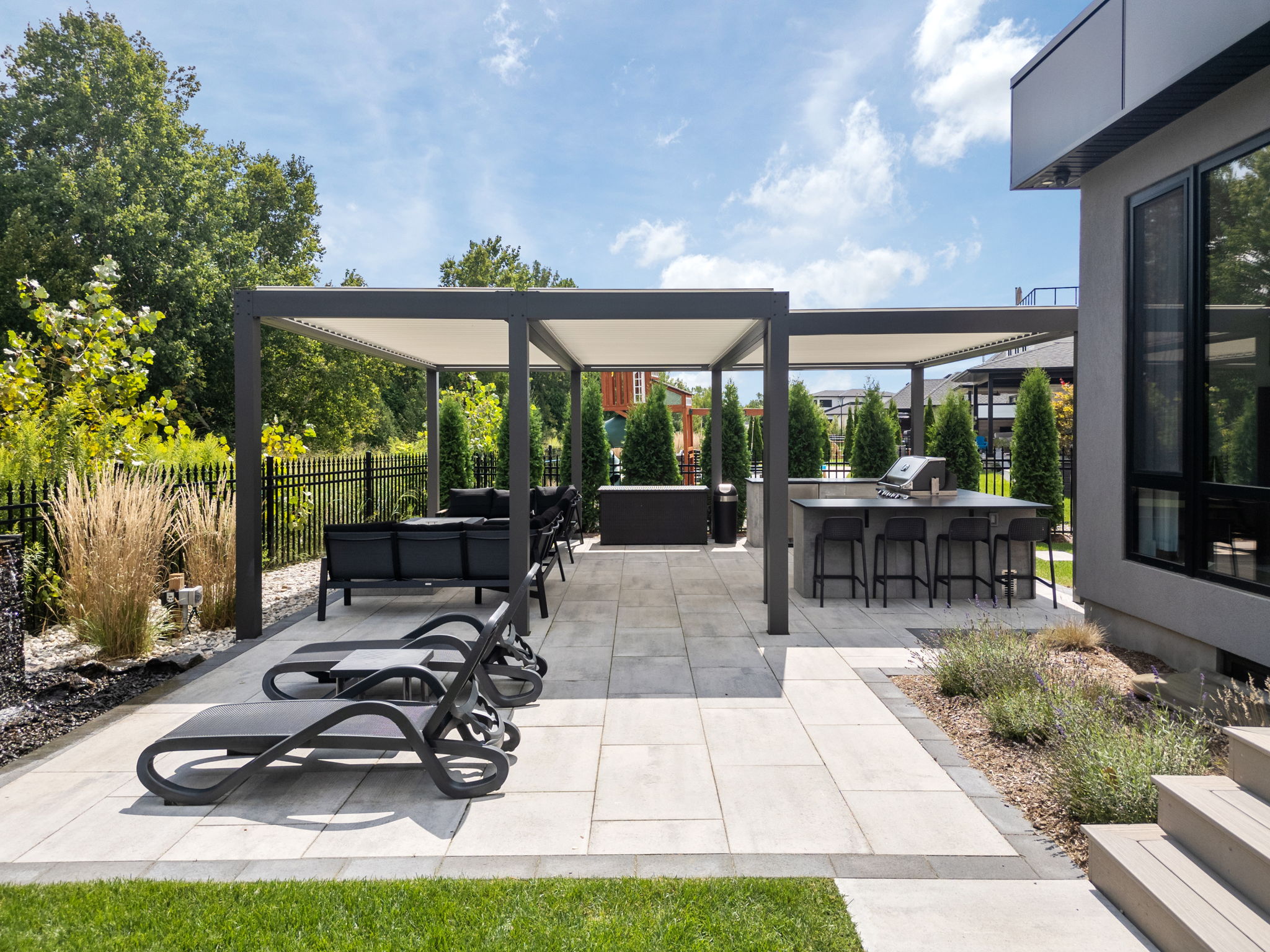 Modern outdoor patio with pergola, lounge chairs, and dining area. Surrounded by greenery and equipped with a grill. Spacious and inviting atmosphere.