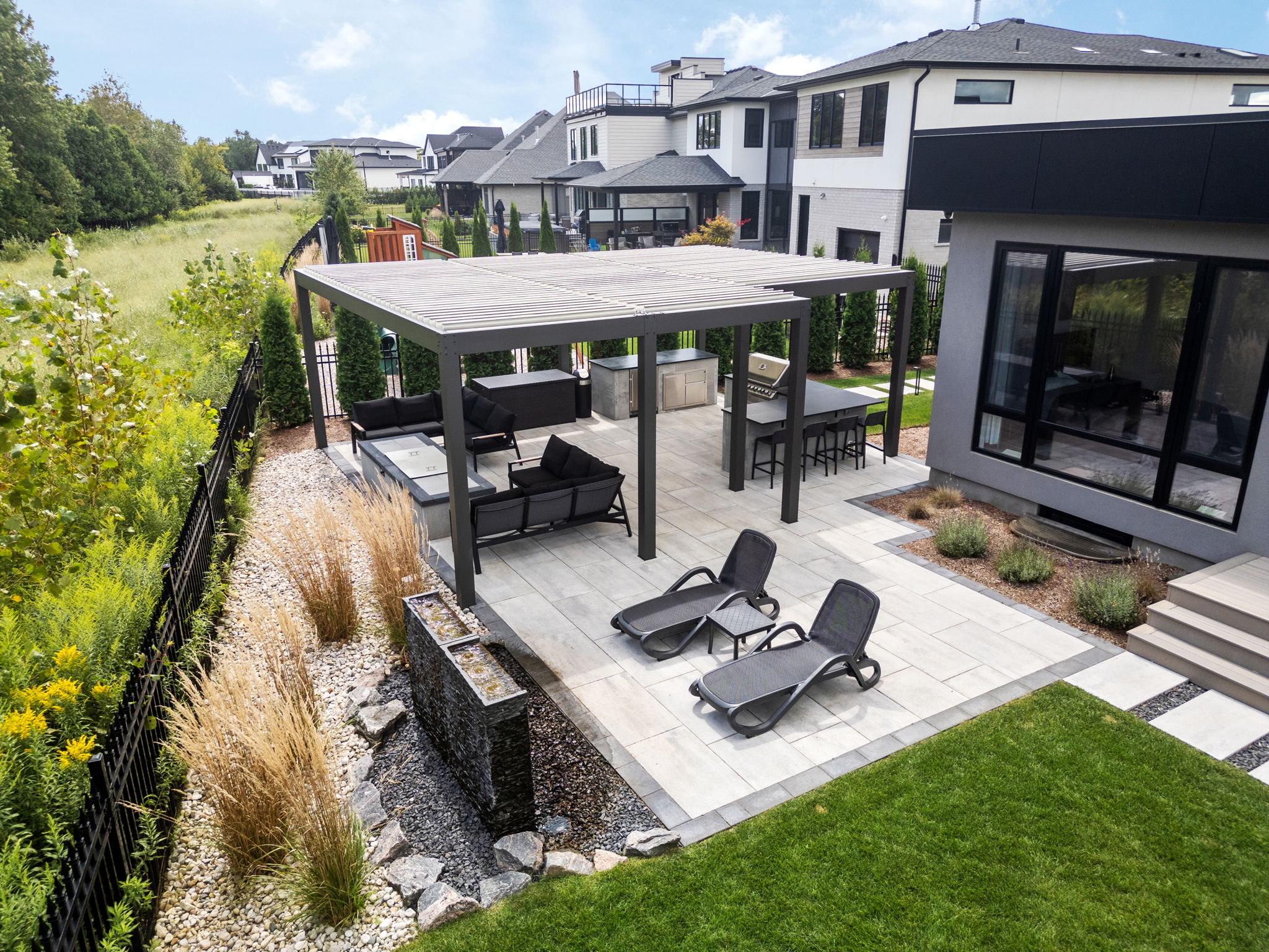 Modern patio with lounge chairs, pergola, and seating area. Surrounded by lush greenery and contemporary houses, creating a serene backyard atmosphere.