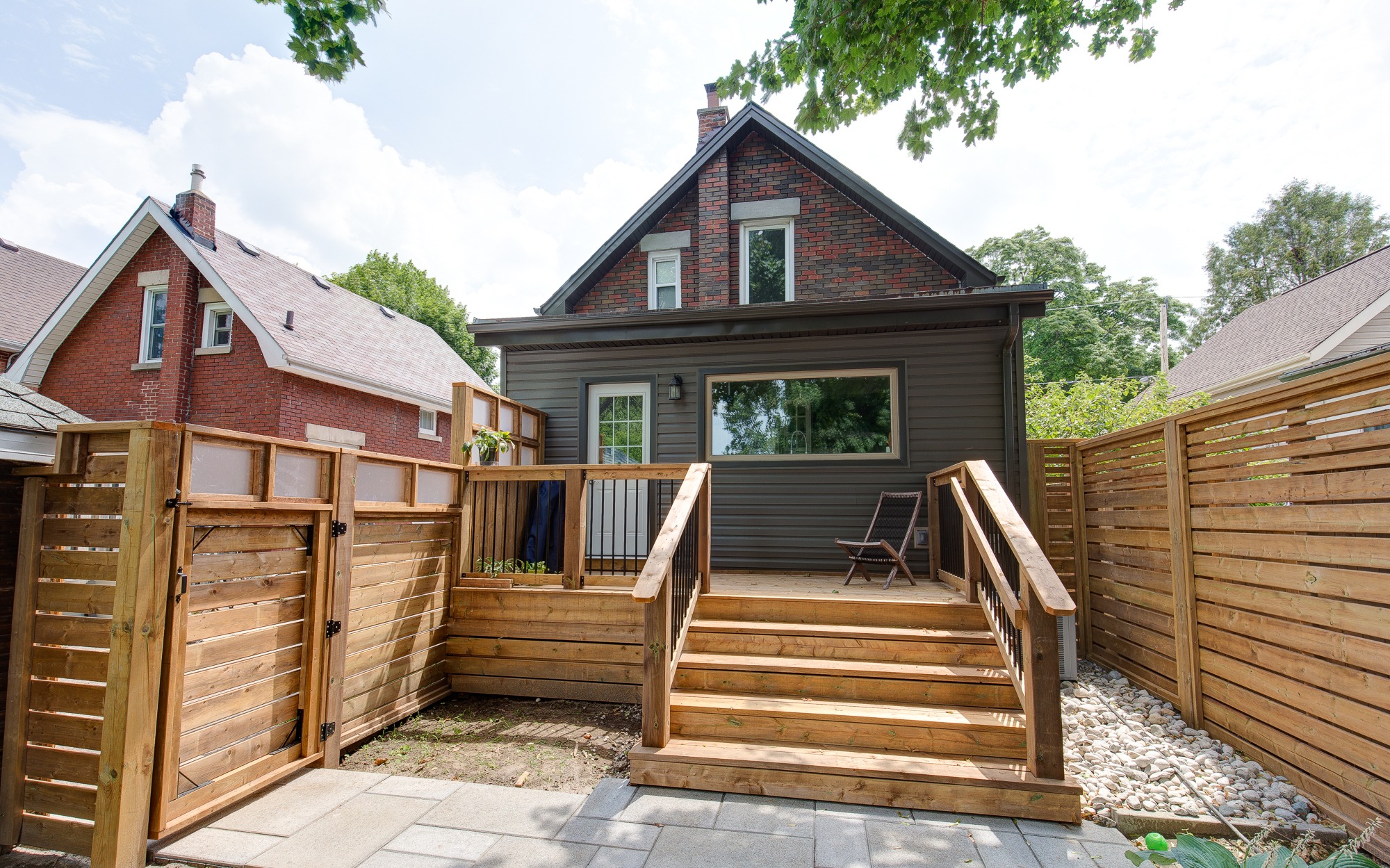 A charming brick house with a spacious, wooden deck and fenced yard, surrounded by lush greenery under a partly cloudy sky.