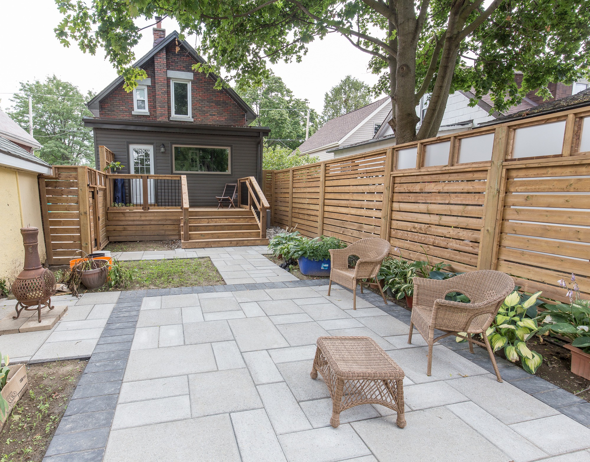 A backyard patio with wicker furniture, surrounded by wooden fencing, trees, and plants. A small house with a deck is visible.