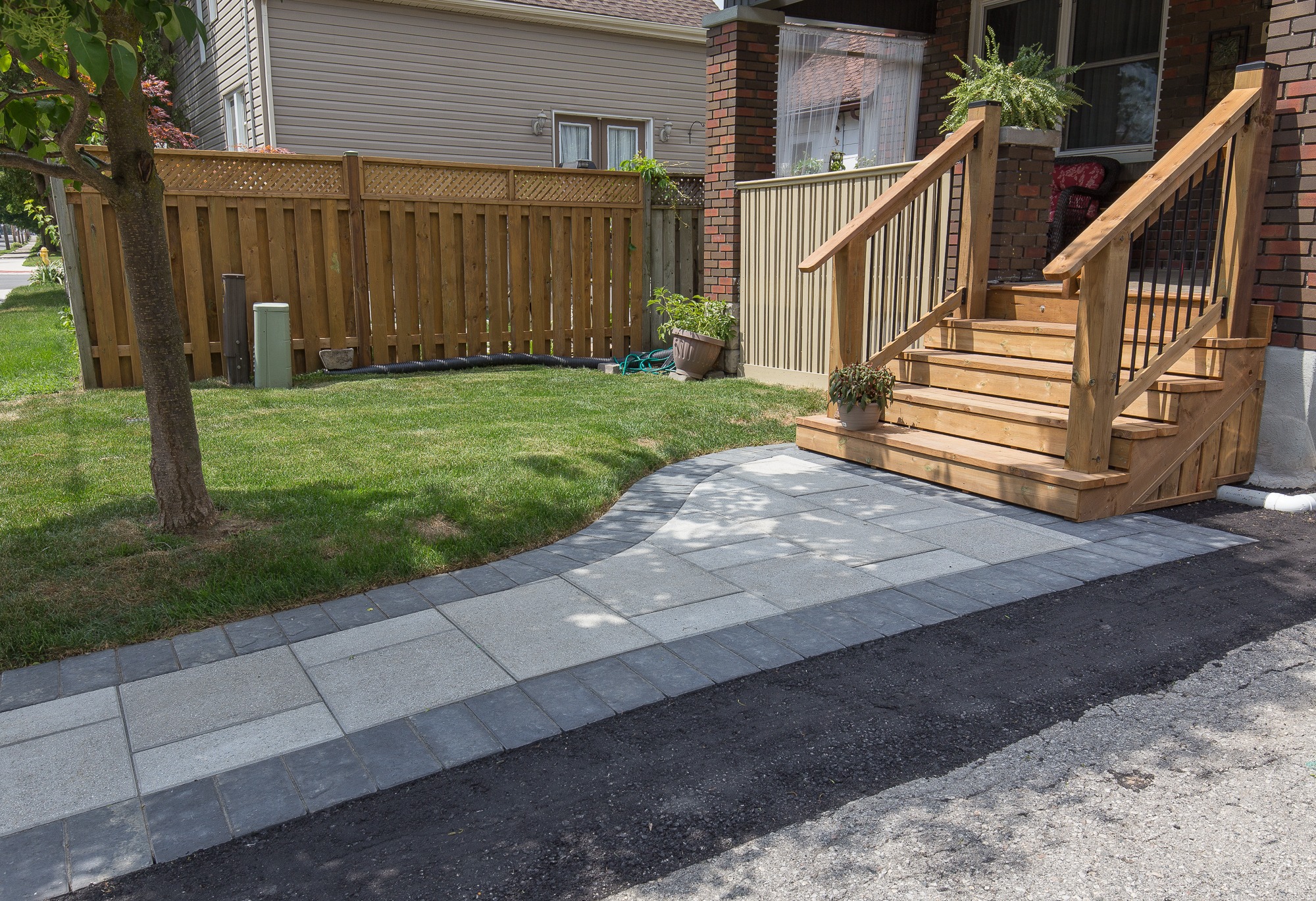 A wooden porch with potted plants and steps leads to a paved walkway beside a grassy lawn and wooden fence. No landmarks visible.