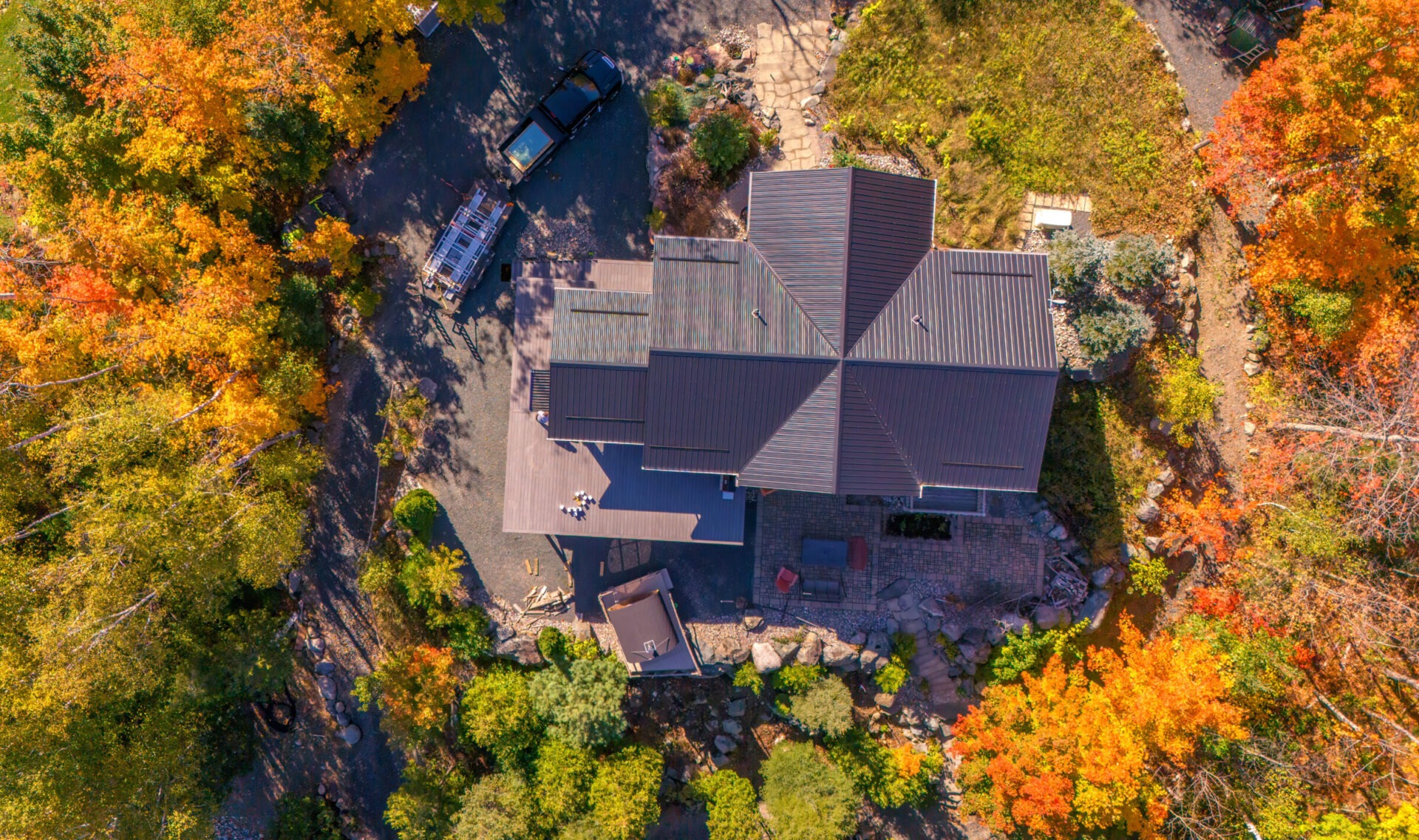 Aerial view of a house surrounded by vibrant autumn foliage, cars parked nearby, and a pathway through the garden. No recognizable landmarks visible.