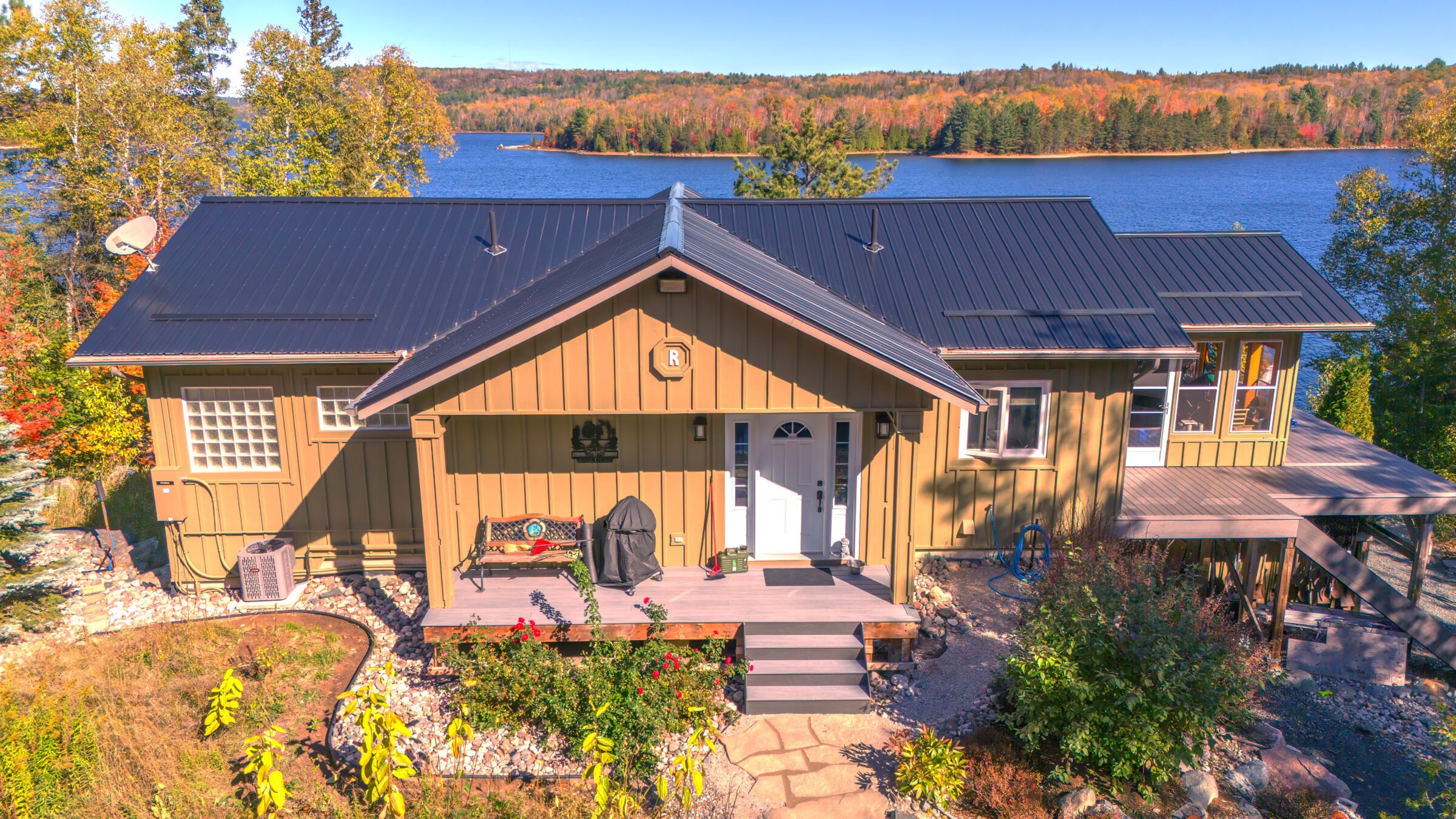 A cabin with a dark roof sits by a serene blue lake, surrounded by colorful autumn trees and a clear sky.