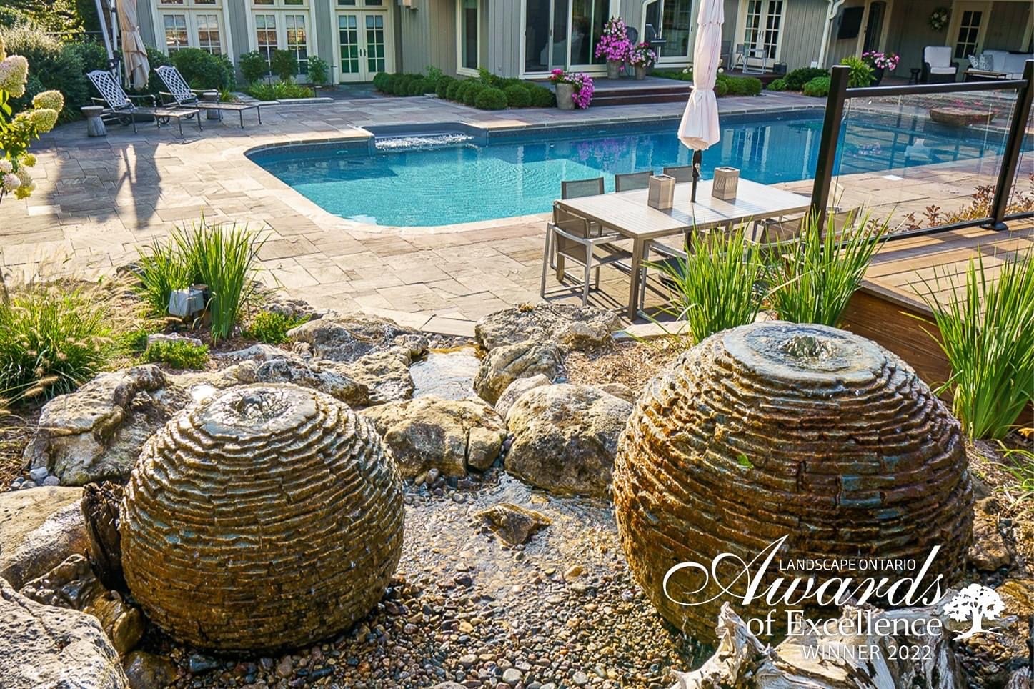 Backyard scene with pool, patio furniture, and decorative stone fountains. Lush greenery surrounds the area, creating a serene and inviting atmosphere.