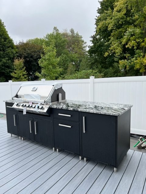 A modern outdoor grill kitchen setup on a gray deck, surrounded by white fencing and lush green trees, under an overcast sky.