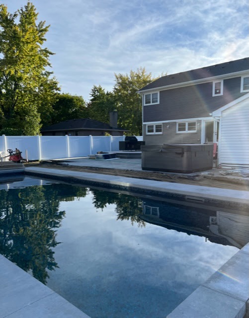 A backyard features a pool, a jacuzzi, and a house with a fenced perimeter. Trees are visible under a clear sky.