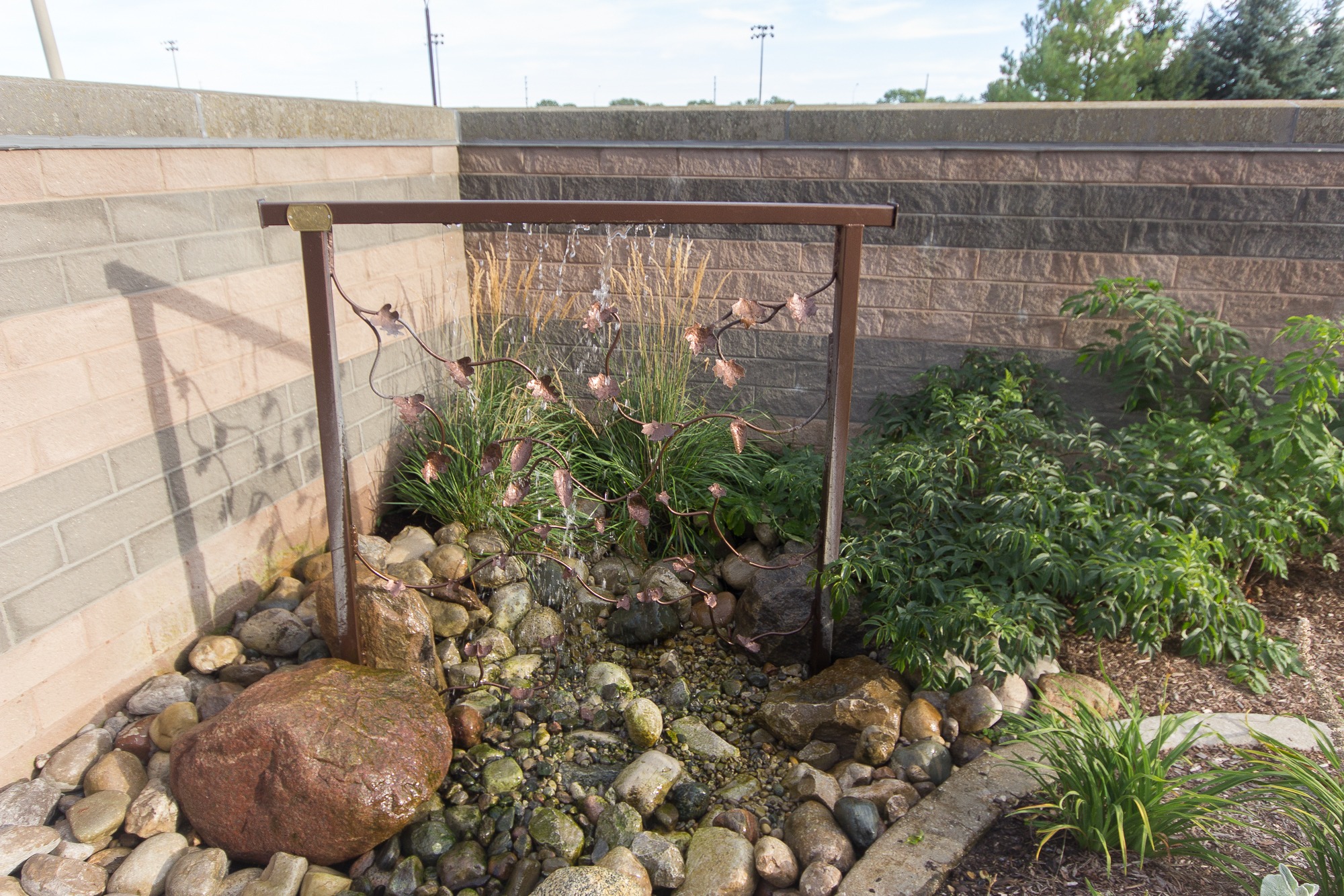 A small, decorative garden fountain with rocks, plants, and a metal trellis. It's set against a brick wall, enhancing natural beauty.