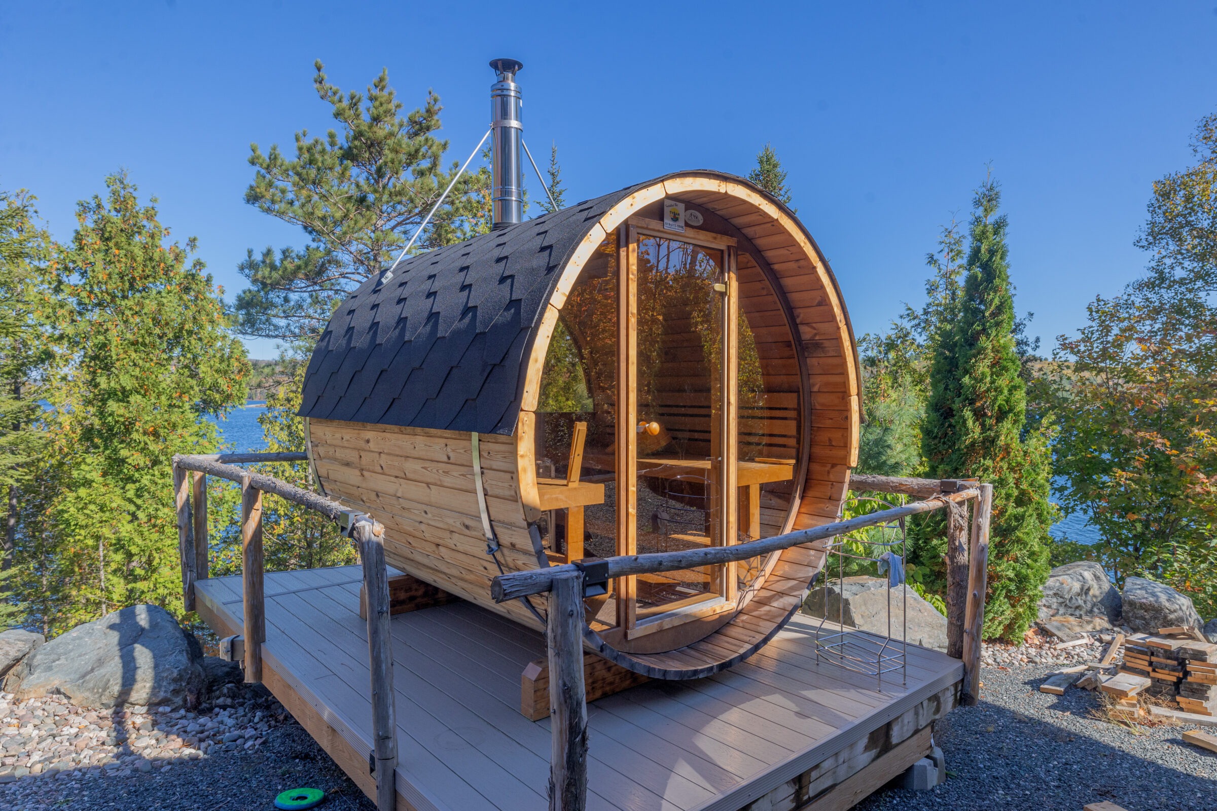 A wooden barrel sauna with glass doors on a deck overlooks a serene lake, surrounded by trees on a clear, sunny day.