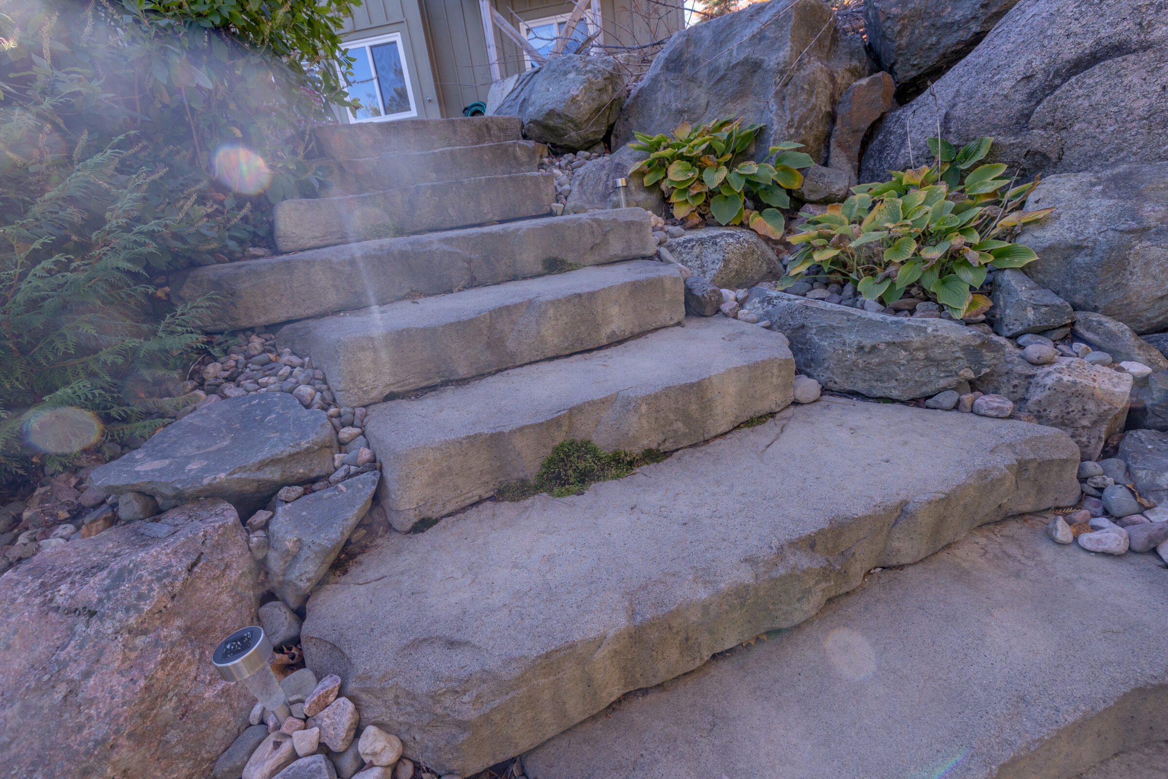 Stone steps surrounded by rocks and greenery lead to a house. Sunlight creates a lens flare, adding a dreamy effect.
