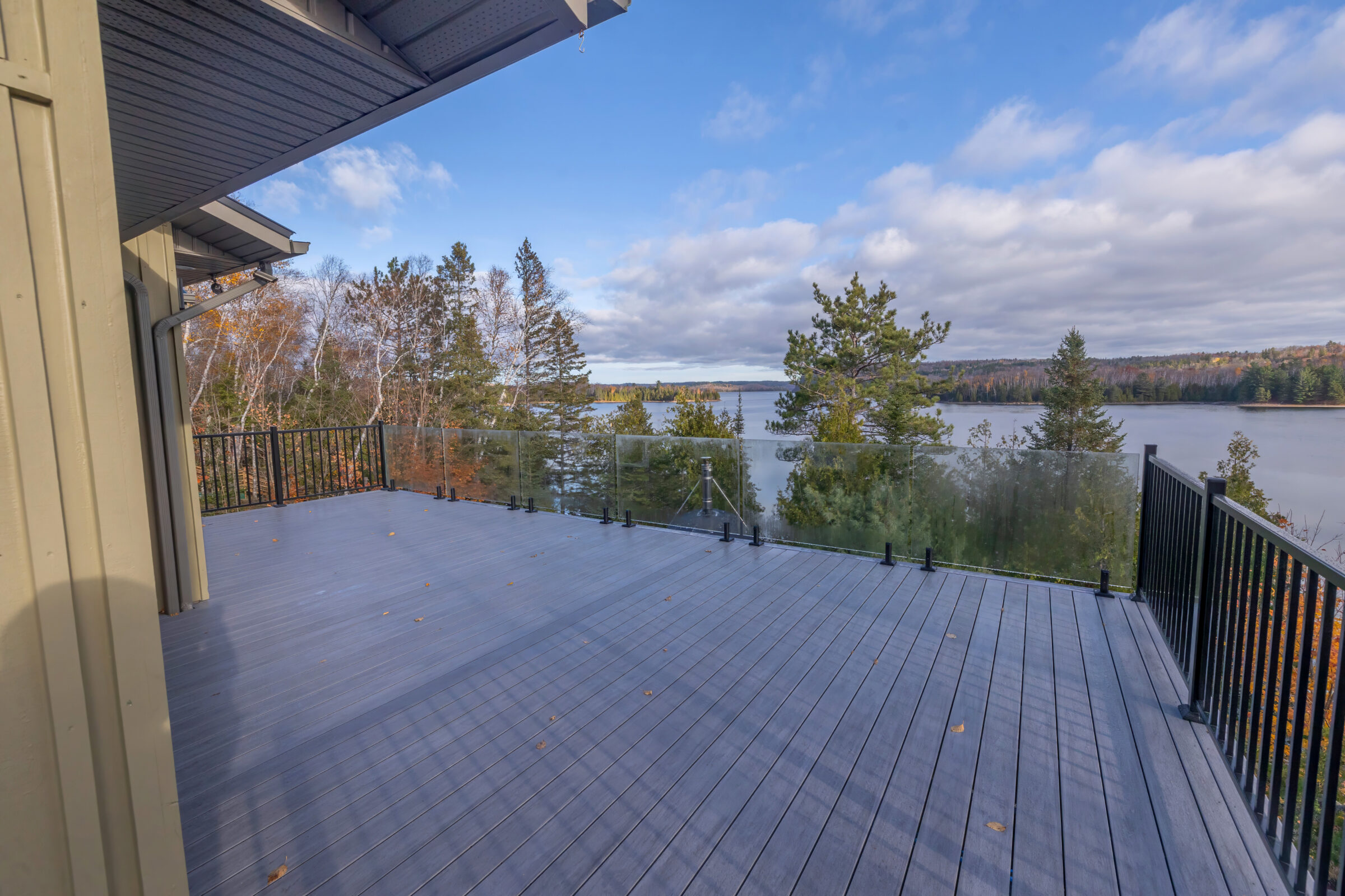 Spacious wooden deck with glass railing overlooks scenic lake and forest under a clear blue sky, creating a serene natural atmosphere.