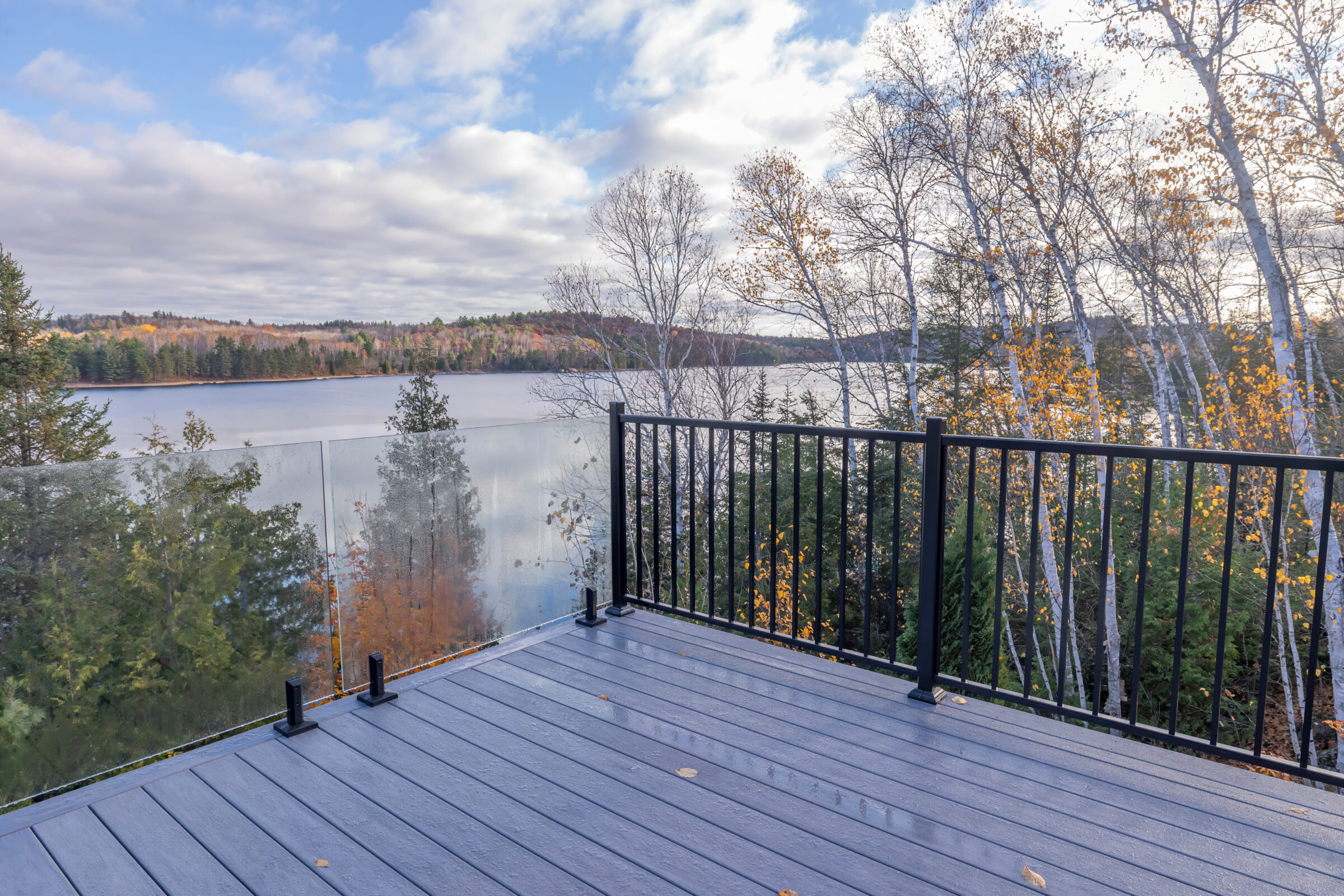 A deck overlooks a serene lake surrounded by autumn trees. The sky is partly cloudy, creating a peaceful, picturesque atmosphere.