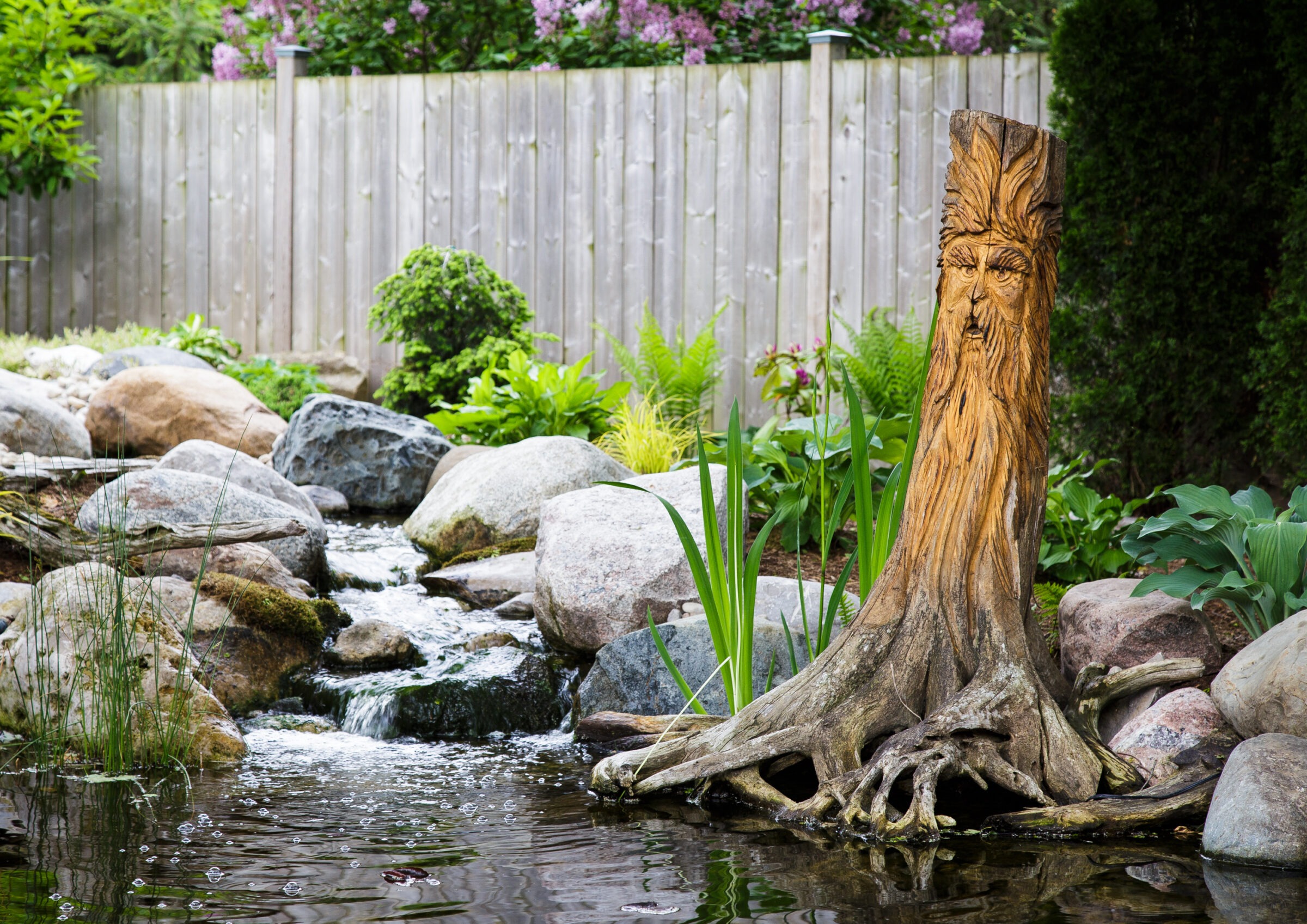 A wooden sculpture of a face stands by a small garden stream, surrounded by rocks and vibrant green plants, under a wooden fence.