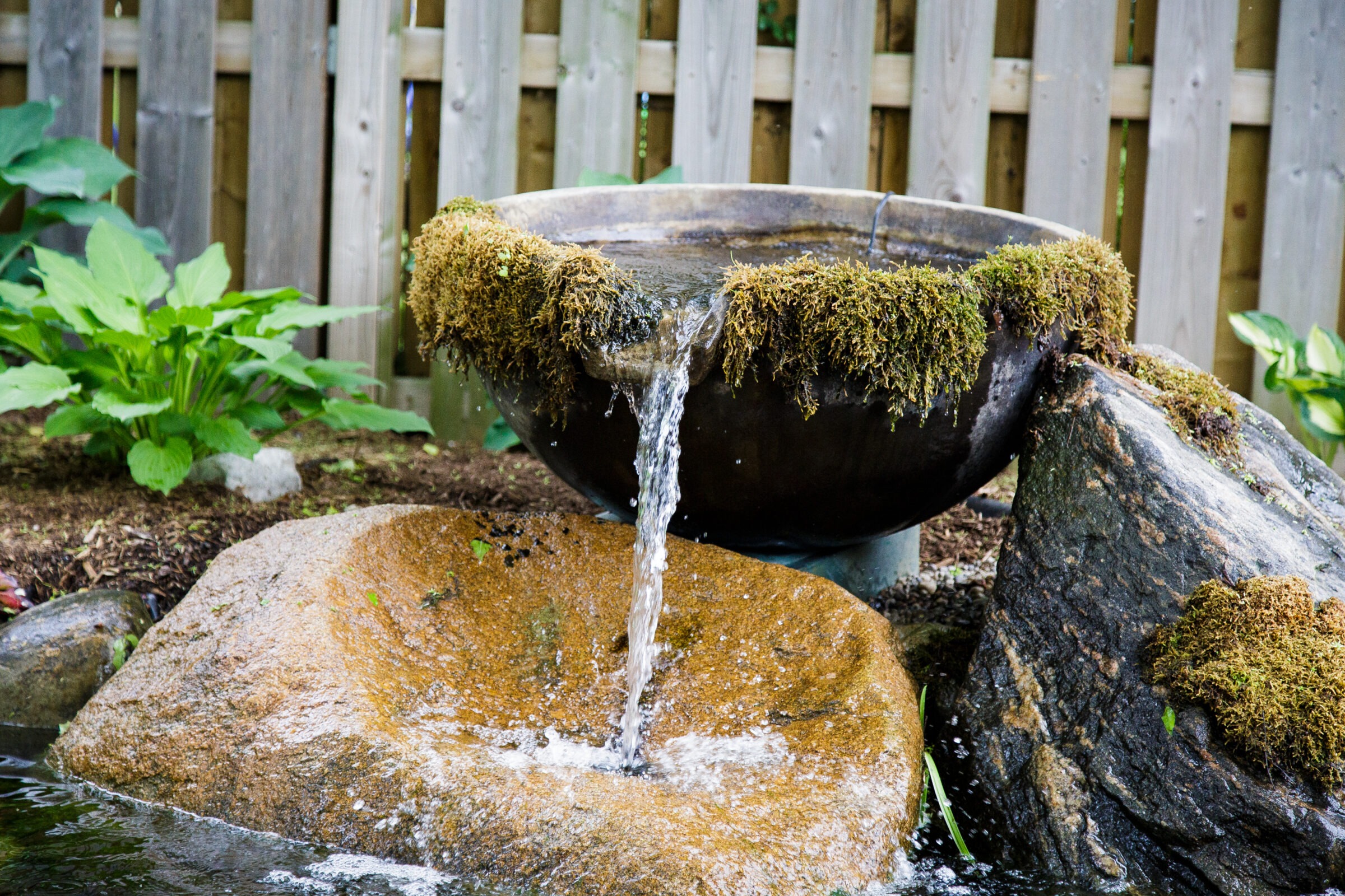A rustic garden fountain with moss-covered bowl spilling water into a stone basin, surrounded by greenery and a wooden fence backdrop.