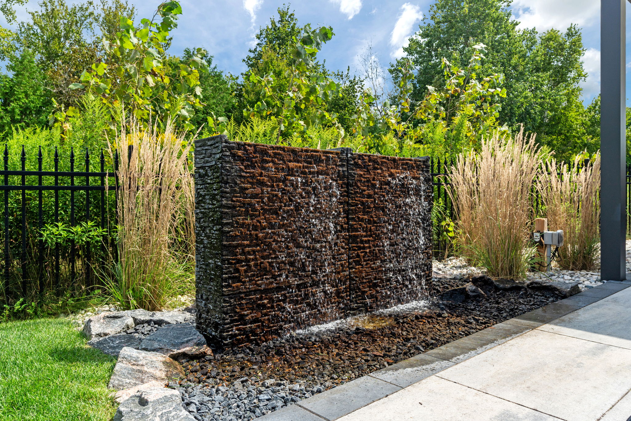 Brick water feature pours over rocks in a landscaped garden, surrounded by tall grasses and trees under a clear blue sky.