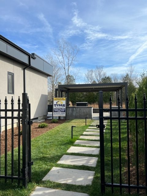 A modern backyard with a pergola, black fence, stone path, and a sign for Tydan Contracting. Trees and blue sky in the background.