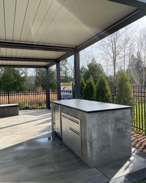 Outdoor kitchen under modern pergola with sleek countertop and cabinets. Surrounded by trees and a fence, with a "Tydan" sign visible.
