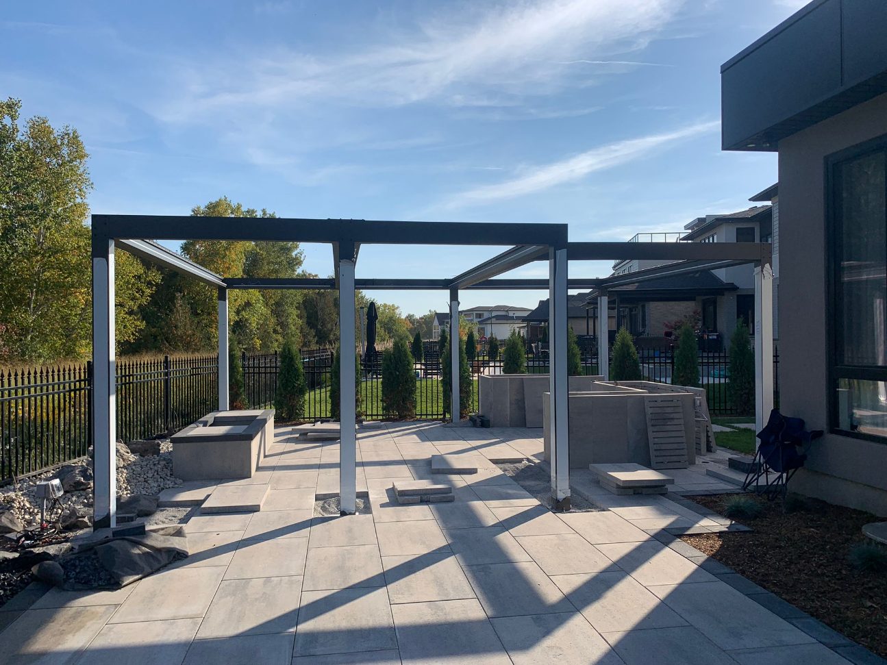 A modern backyard is under construction with a pergola, patio, and landscaped garden, surrounded by a fence and neighboring houses.