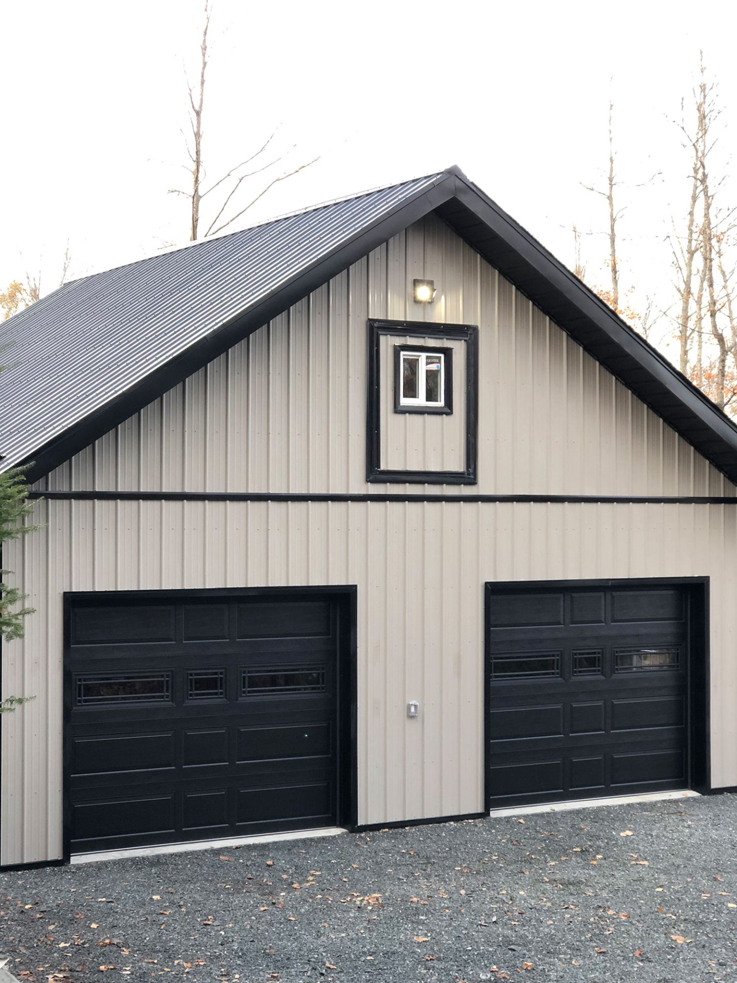 A beige metal garage with a black roof, two black garage doors, small window, set against a backdrop of leafless trees.