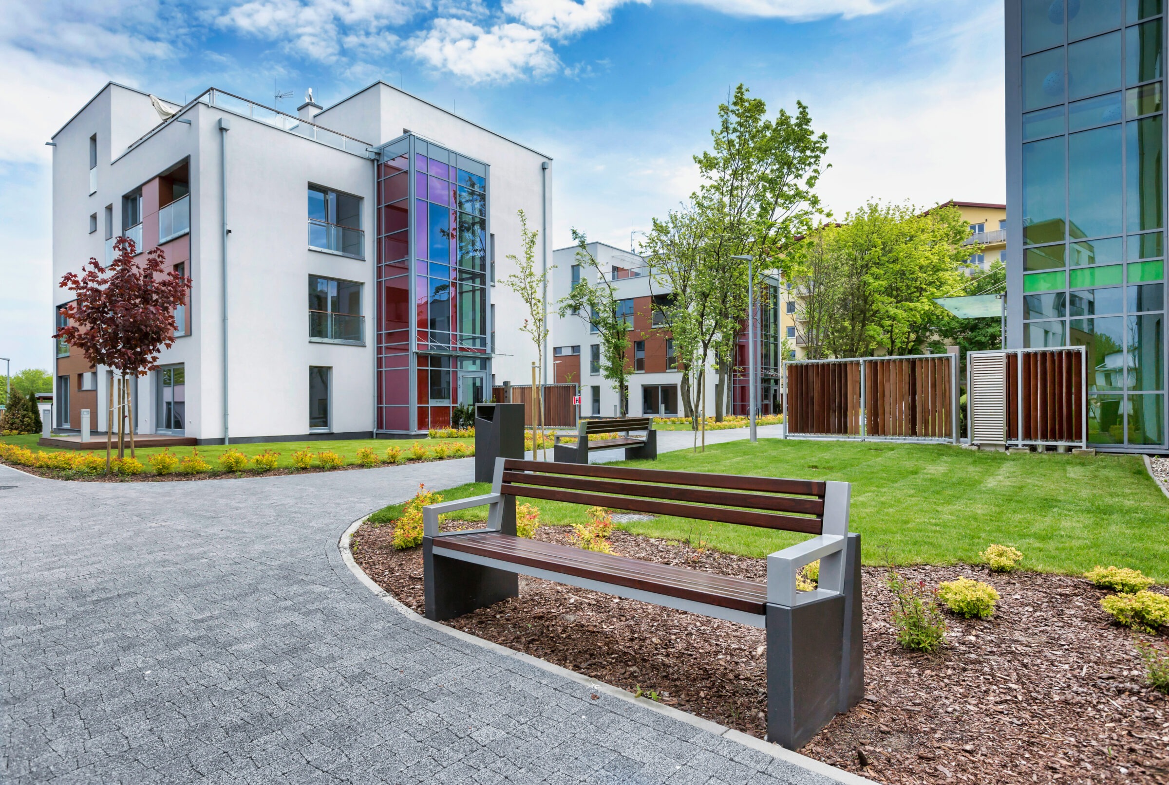 Modern apartment buildings with colorful glass facades surrounded by greenery and paths. Benches and small trees line the landscaped area under a blue sky.