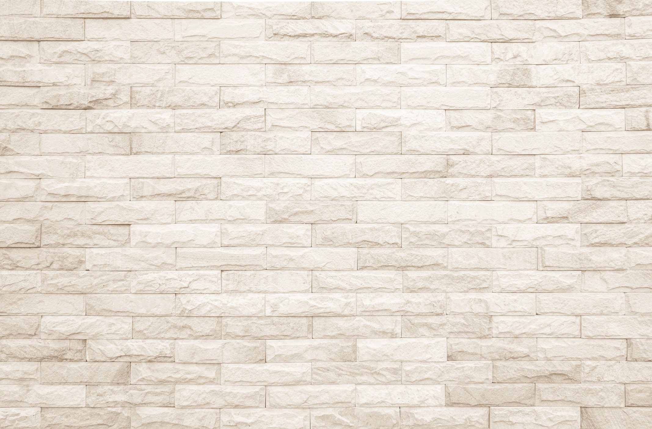 A person stands against a textured stone wall, wearing a black coat and blue jeans. Sunlight casts subtle shadows on the bricks.